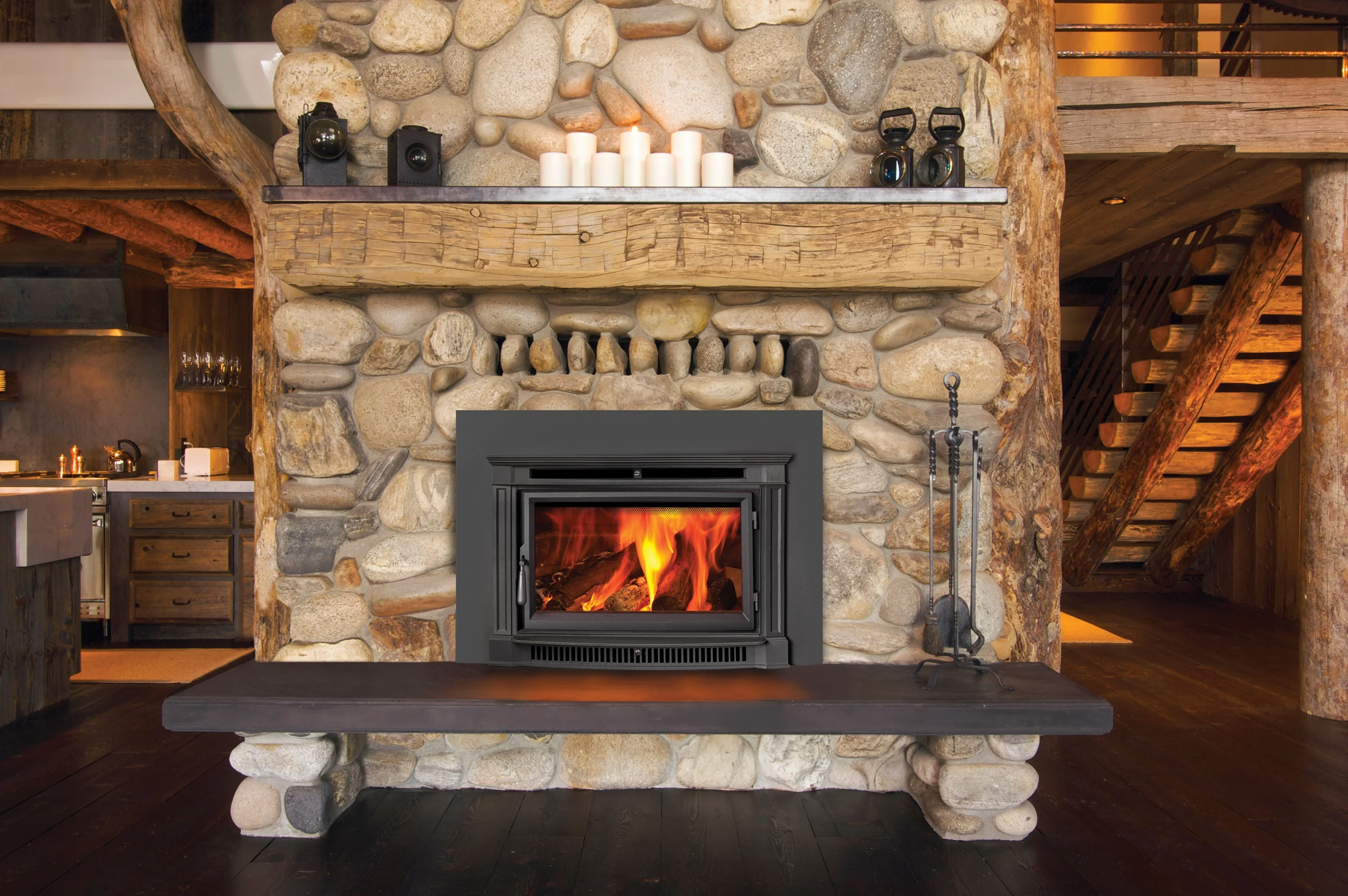 A rustic stone fireplace with a wooden mantel is lit, showing flames. On the mantel are white candles and lanterns. The surrounding area has a wooden interior with stairs and a kitchen visible.