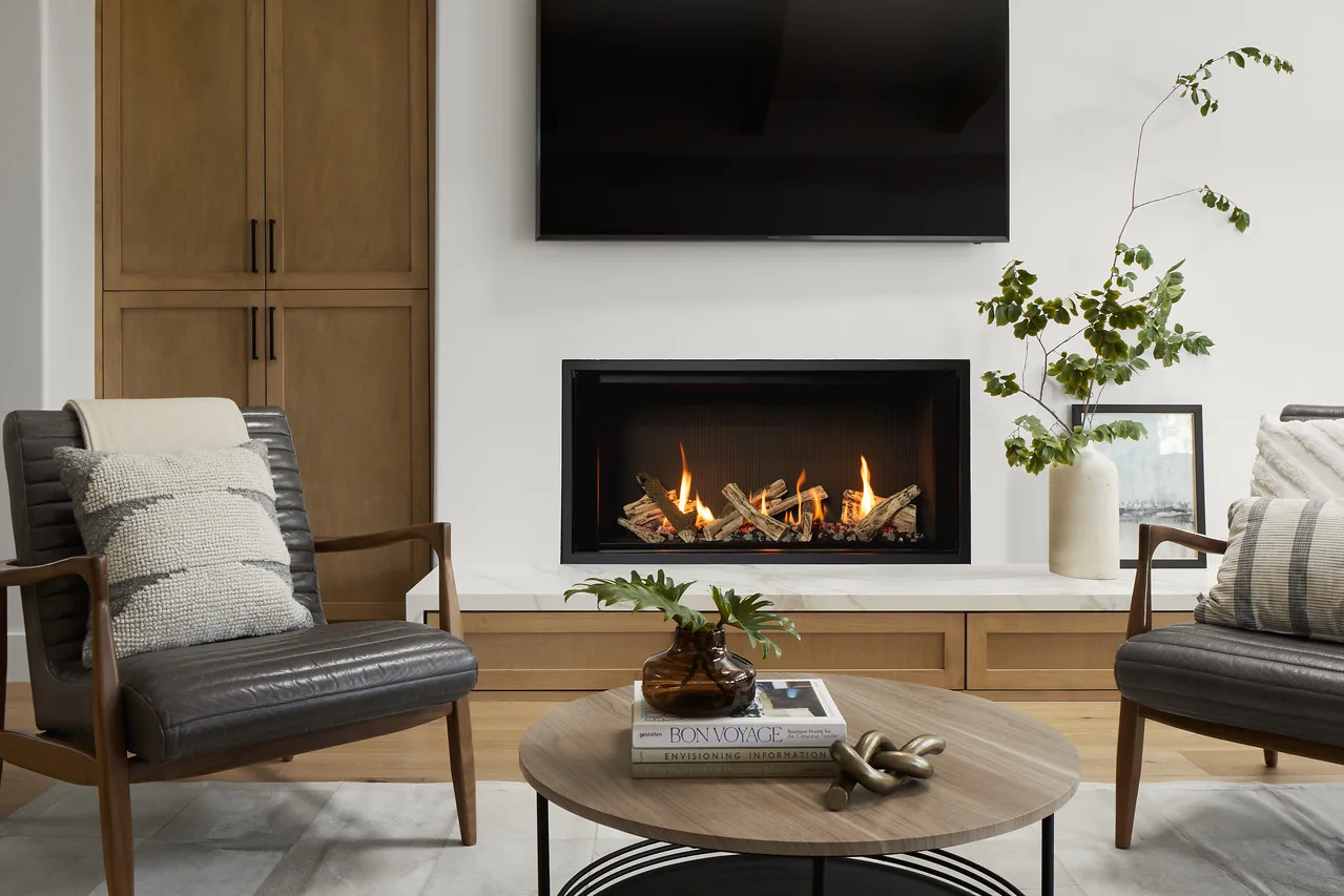 Modern living room with gas fireplace under a wall-mounted TV, wood cabinets, two leather armchairs, round coffee table with books and vase, and a tall leafy branch.