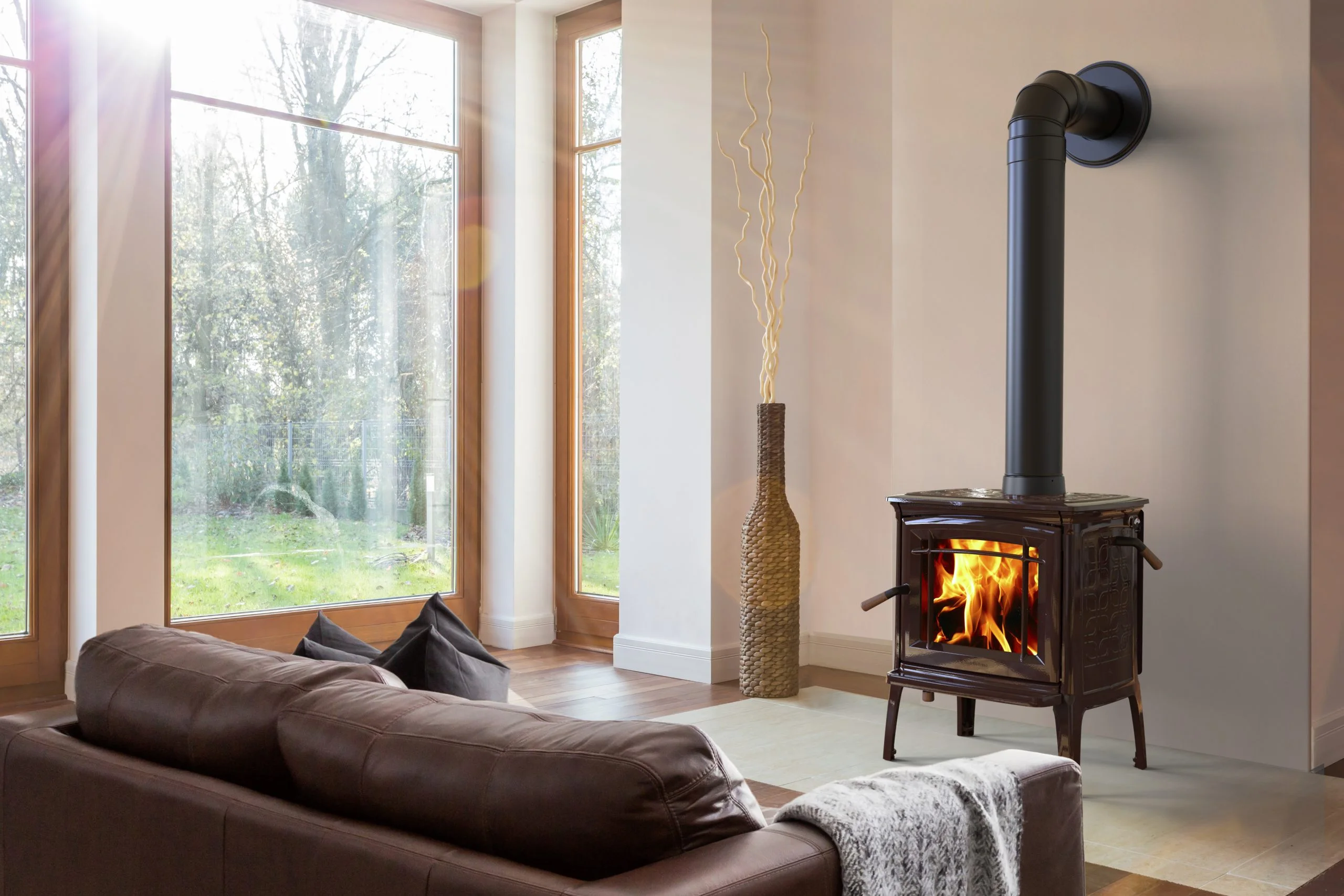 A bright living room featuring a black freestanding wood-burning stove with a visible fire, situated next to large windows looking out at a green landscape. A brown leather sofa sits in the foreground, and a tall floor vase with decorative yellow branches stands in the corner.