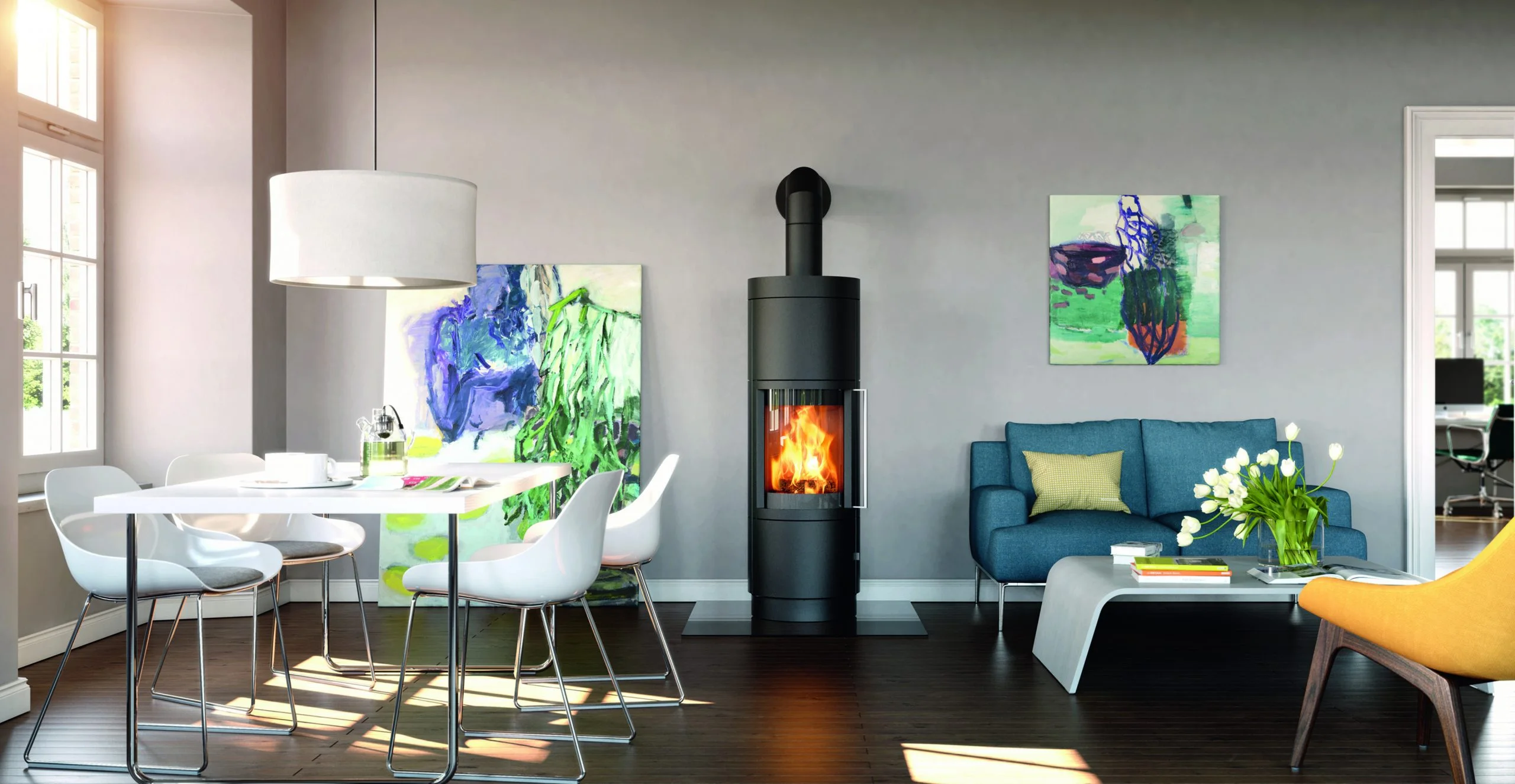 A brightly lit, modern living room and dining area featuring a tall, black, cylindrical freestanding wood stove with a visible fire. The stylish space includes a white dining table with chairs, a blue sofa, a yellow lounge chair, and vibrant abstract paintings on light grey walls.
