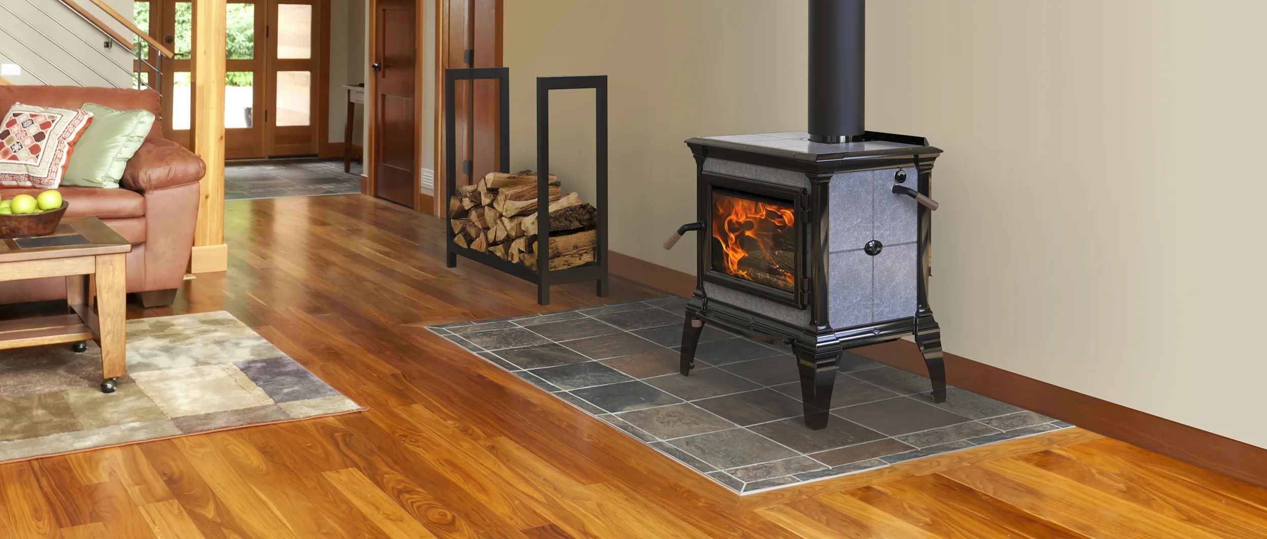 A black freestanding wood-burning stove with a lit fire, sitting on a gray tiled hearth in a room with hardwood floors. Next to the stove is a black metal rack filled with firewood. In the background, a brown leather sofa with patterned pillows sits near a staircase.