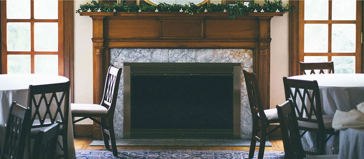 A cozy room with wooden furniture, round tables covered with white tablecloths, and an unlit fireplace with a marble surround. The mantle is decorated with greenery and lit candles.