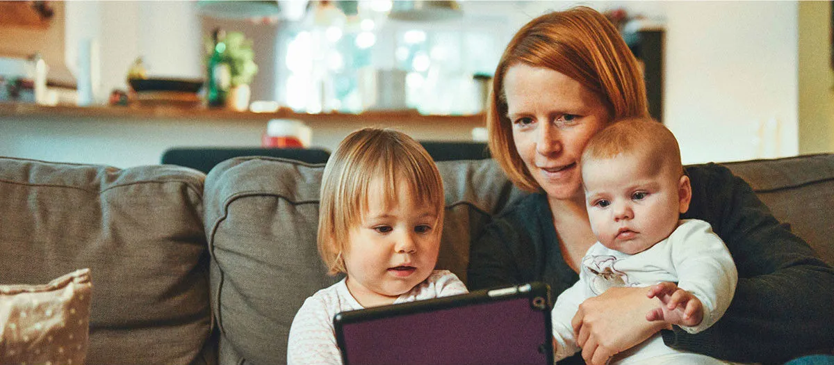 A woman with red hair sits on a couch holding a baby, while a young child sits next to them looking at a tablet. The background shows a blurred kitchen setting.