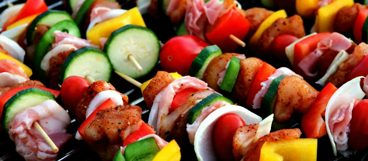 Colorful skewers with grilled meat, bell peppers, zucchini, onion, cherry tomatoes, and bacon arranged closely together on a barbecue grill.