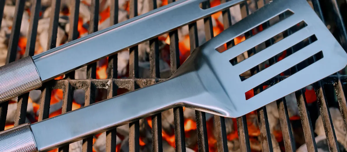 Two stainless steel grilling tools, a fork and a slotted spatula, rest on a hot BBQ grill with glowing charcoal underneath.