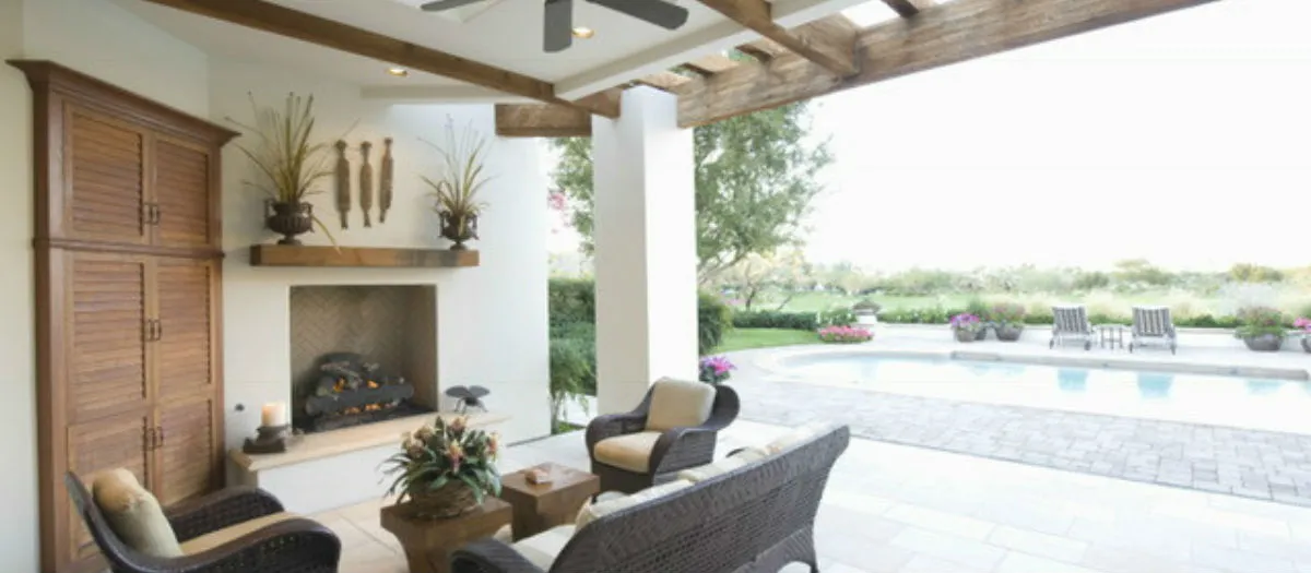 Outdoor patio with wicker furniture, a wooden beam ceiling, and a fireplace with a wooden mantle displaying decorative plants. View of the garden and a pool in the background.