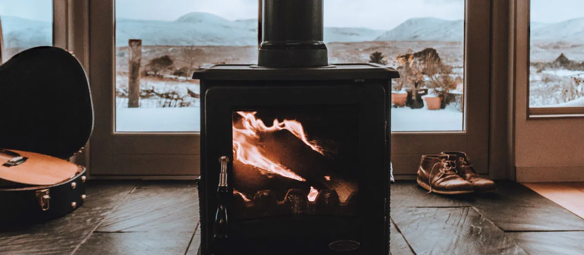 A lit wood-burning stove sits inside a cozy room with a guitar case on one side and a pair of boots on the other. Snow-covered mountains are visible through large windows behind the stove.