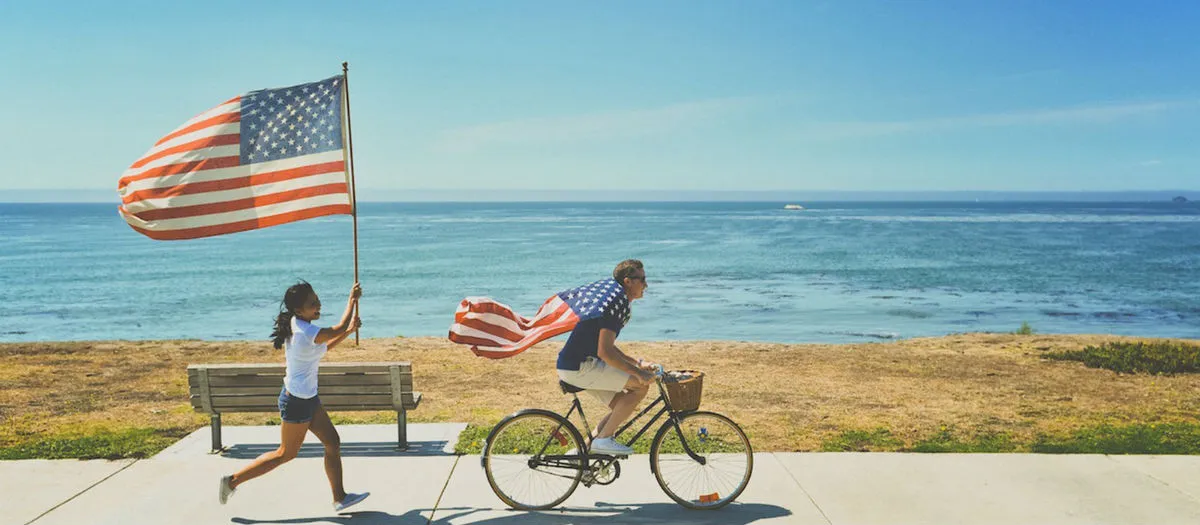 A person rides a bicycle while another person runs behind them holding a large American flag. Both are near the ocean on a sunny day, with a bench and path beside them.