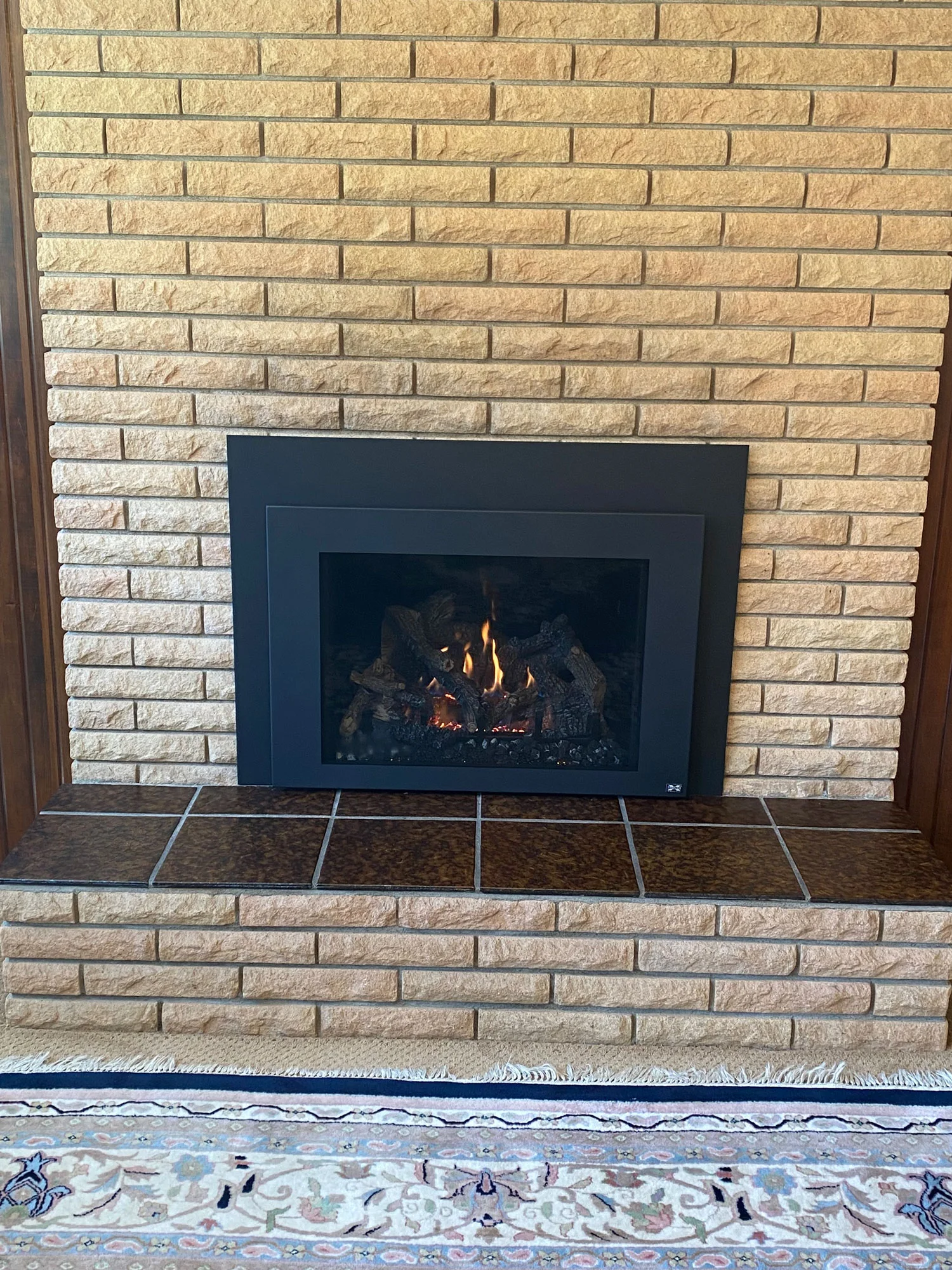 A black-framed fireplace with a burning fire, set against a brick wall. The tiled hearth extends in front, with a patterned rug partly visible at the bottom.