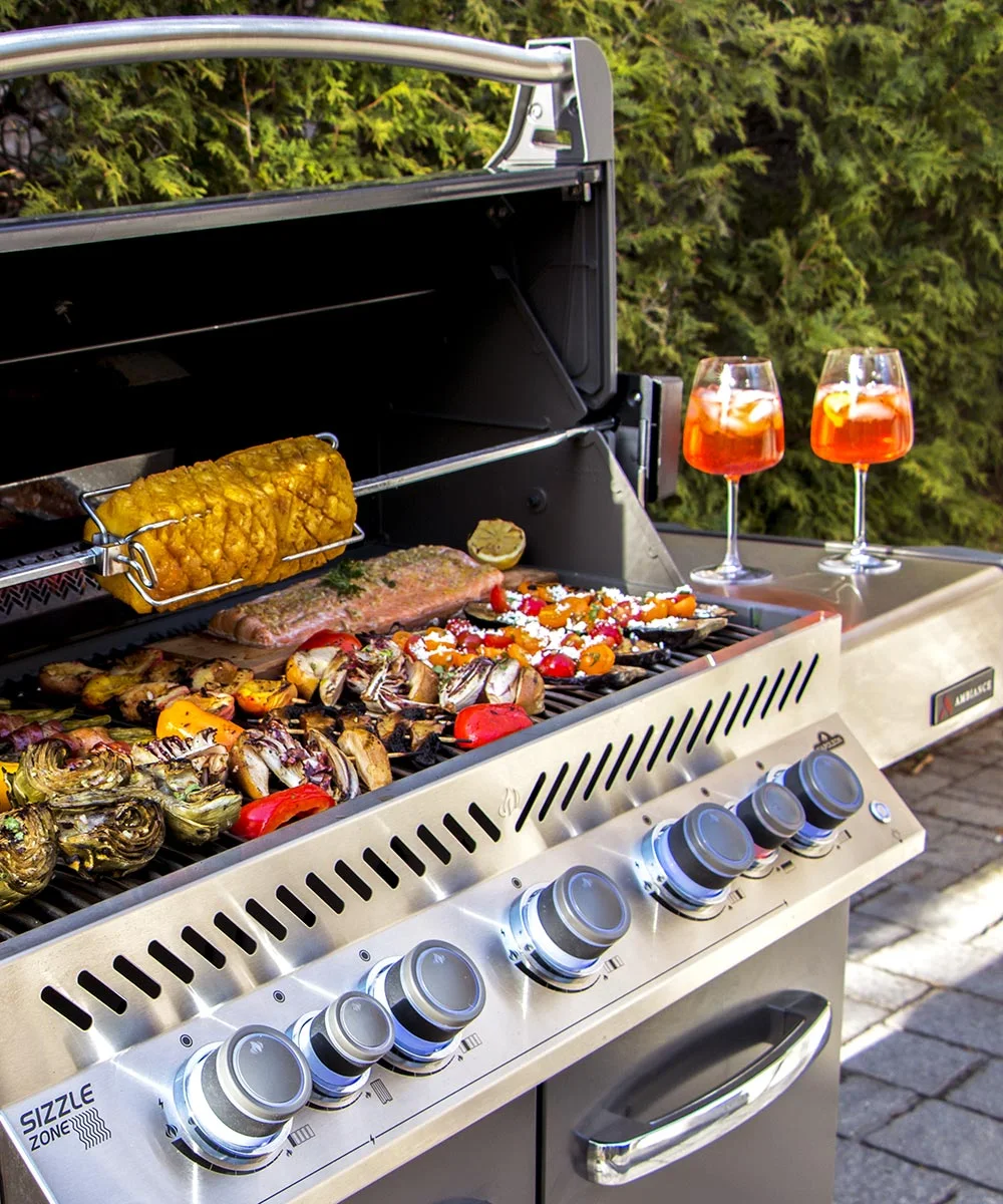 A stainless steel BBQ grill cooking assorted vegetables, corn on the cob, and a large piece of meat, with two orange drinks in glass goblets placed on a nearby surface.