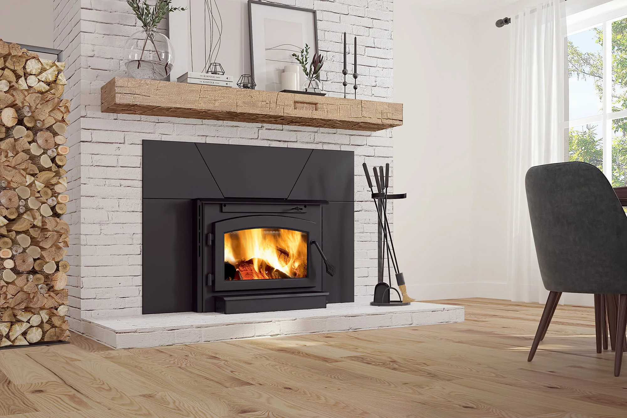 A cozy living room scene featuring a white brick fireplace with a black insert, lit with a fire. A wooden mantel holds decor items. Firewood is stacked to one side, and fireplace tools are on the other.