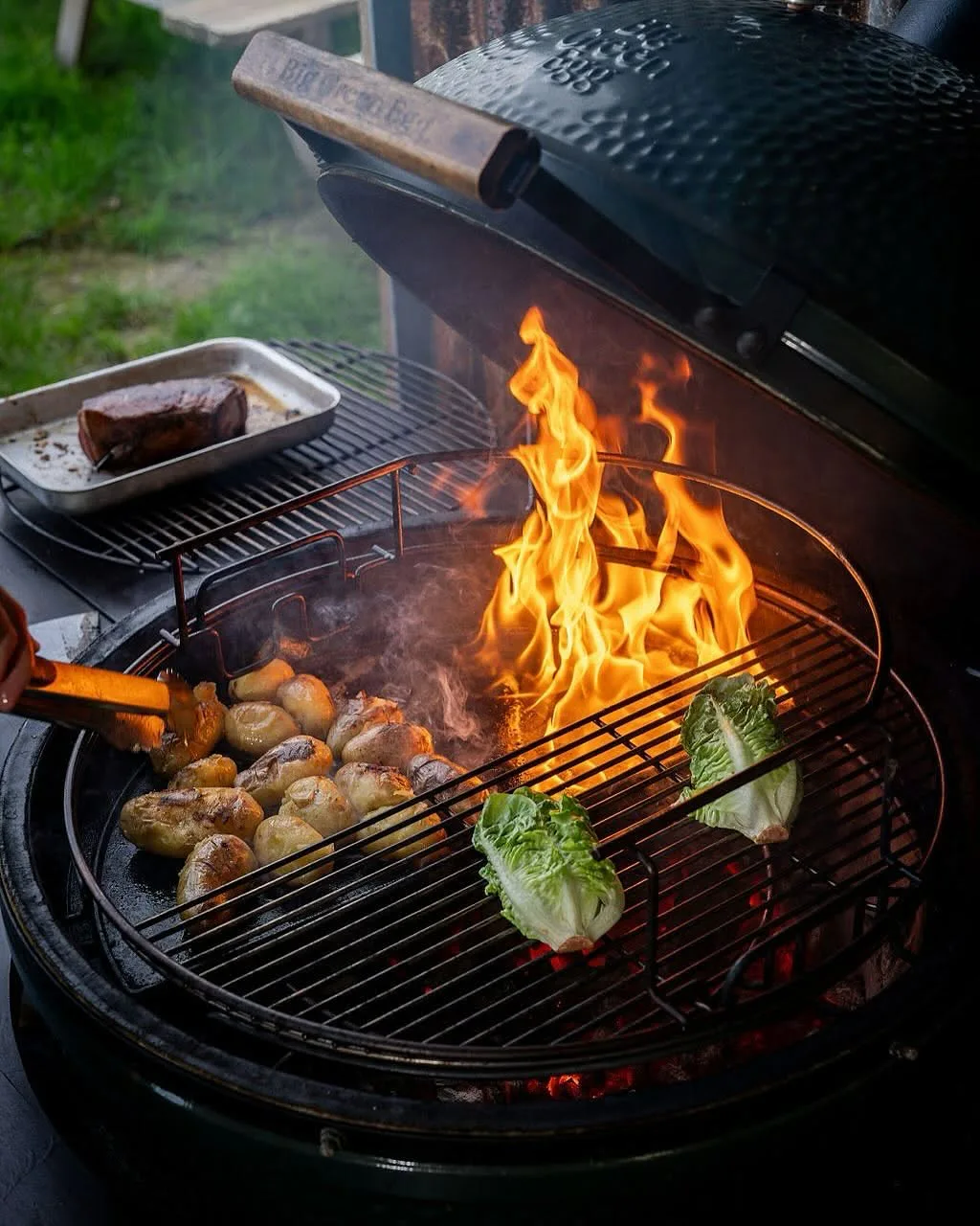 Open charcoal grill with tall flames roasting baby potatoes and halved romaine lettuce while tongs hold a potato