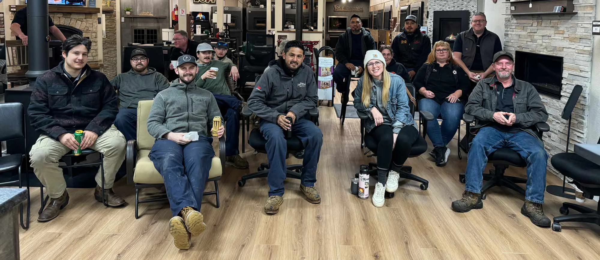 A group of people seated in a fireplace showroom, casually posed and smiling, several holding drinks.