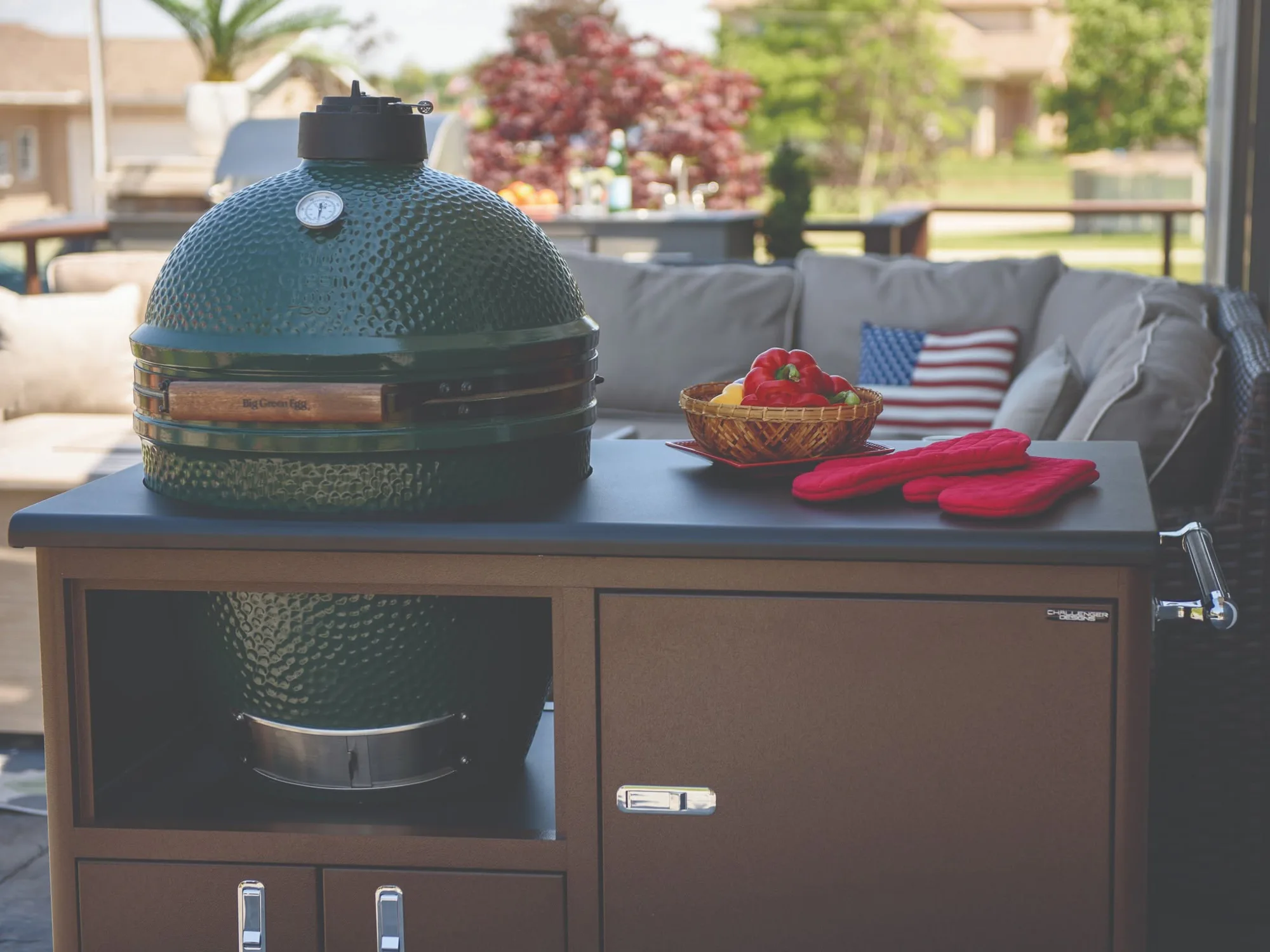 Green kamado grill on a patio cart with basket of bell peppers and red oven mitts, sofa with striped pillow in background