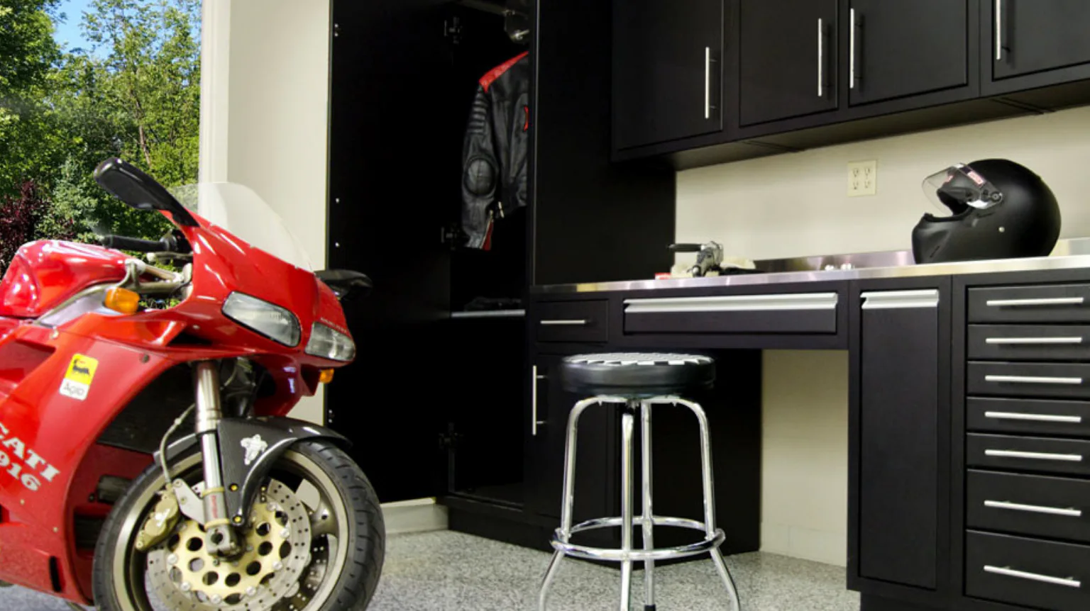 A red motorcycle is parked in a garage featuring black cabinets and a countertop with a motorcycle helmet. A leather jacket hangs open in a nearby closet. A metal stool is nearby.