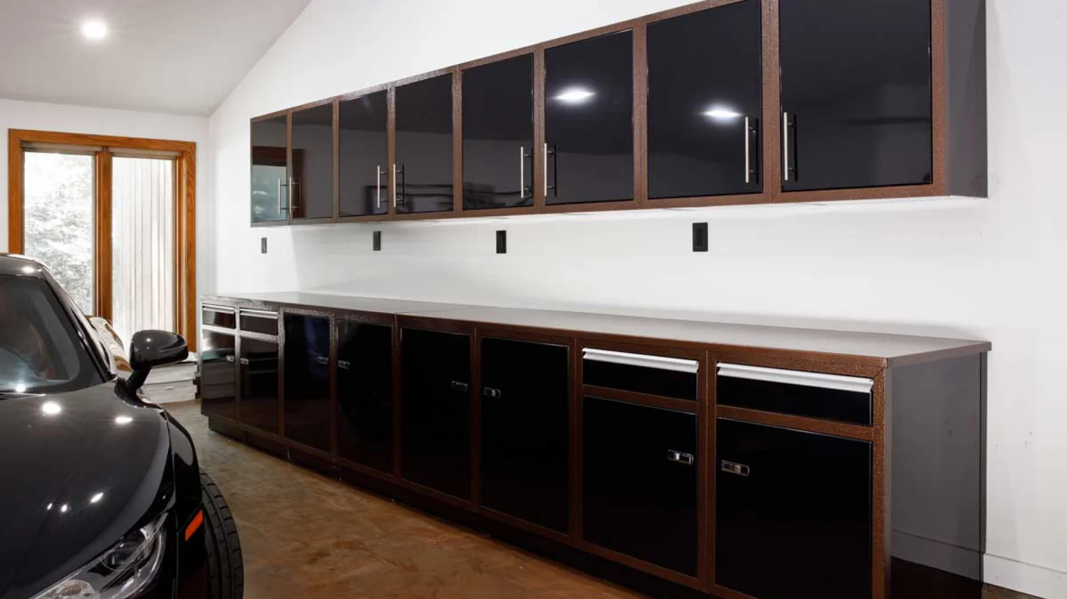 Modern garage with sleek black and wood cabinets, under bright lighting. Cabinets line the walls above and below a smooth countertop. A black car is partially visible in the foreground.