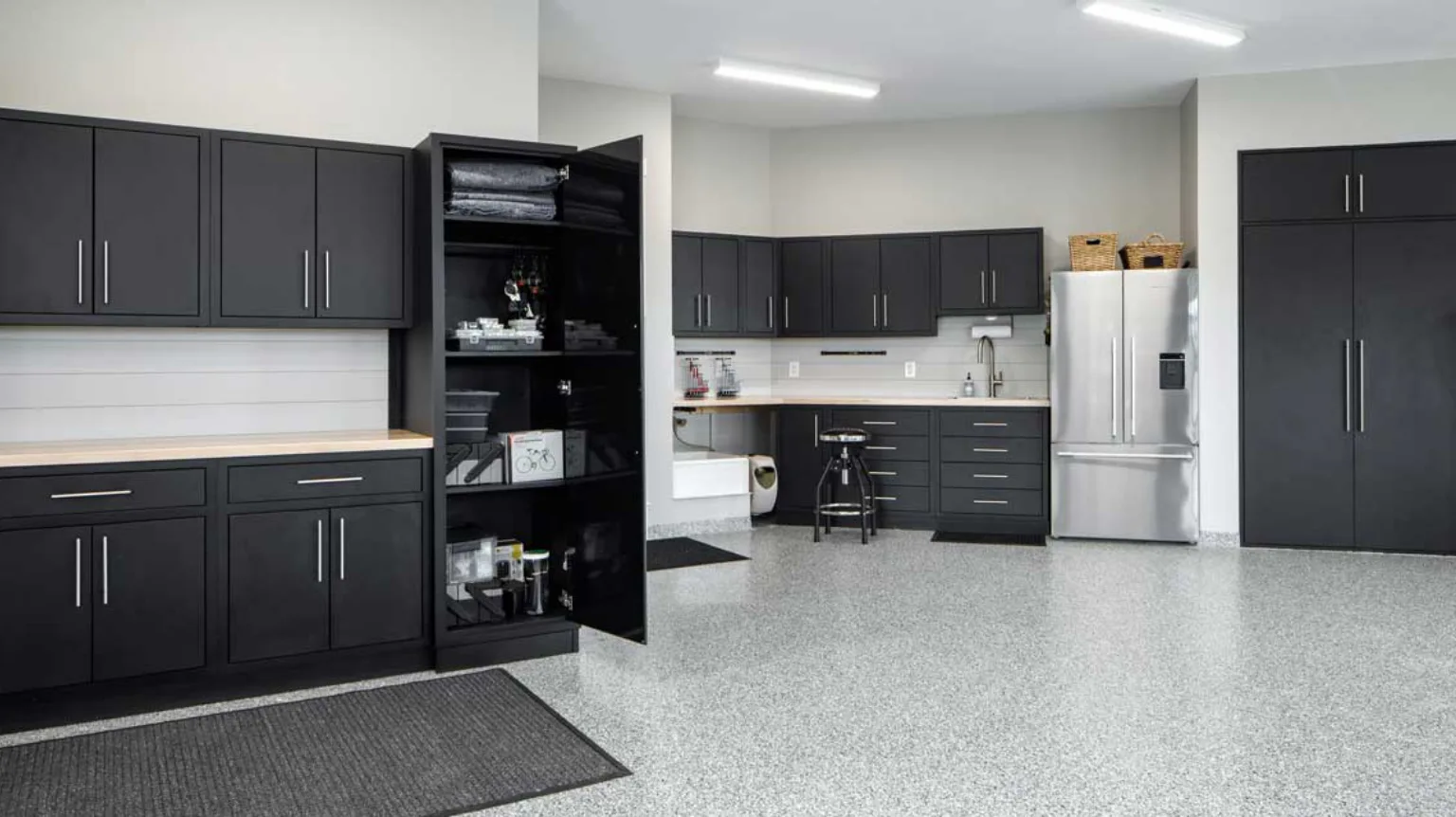 Modern garage interior with dark cabinets, stainless steel fridge, and light countertops. Open cabinet displays tools and supplies. Gray speckled floor and task chair are visible.