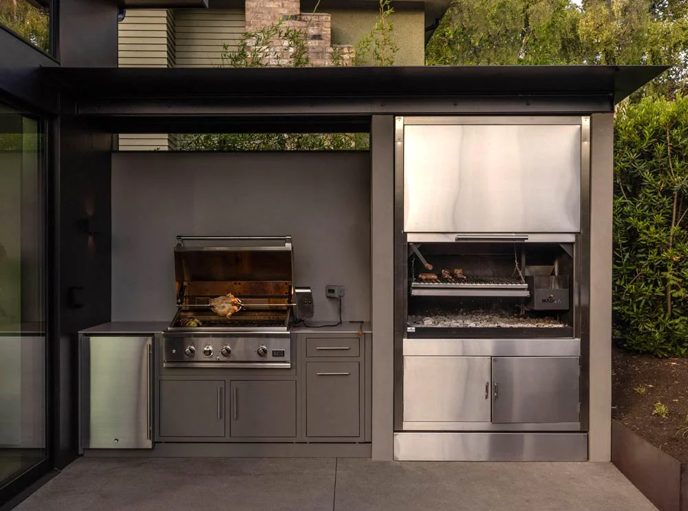 Modern outdoor kitchen with built-in stainless steel gas grill and adjacent charcoal barbecue in gray cabinetry