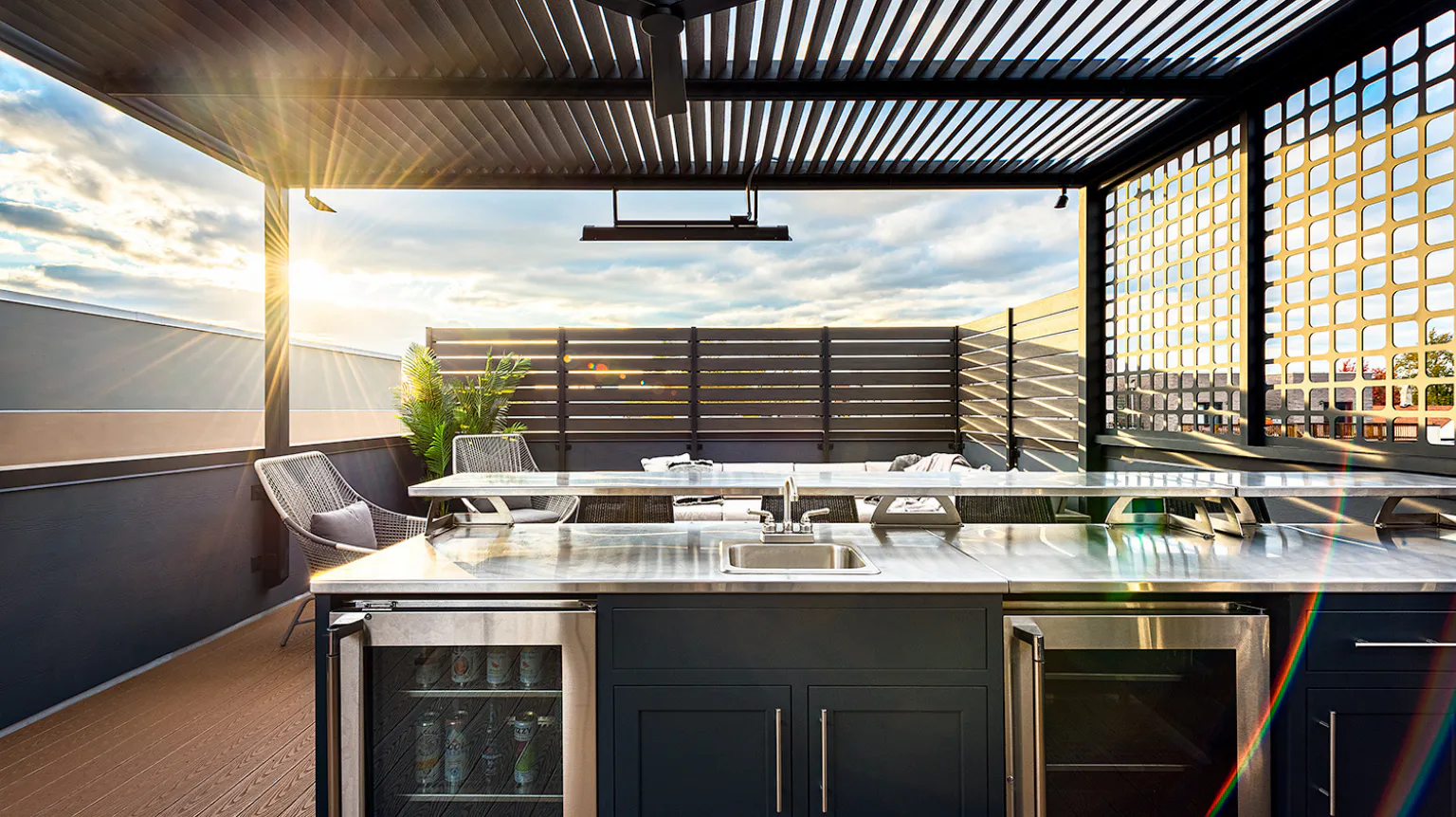 Modern outdoor kitchen with stainless steel counters and open pergola. The sun sets in the background, casting warm light on the seating area with woven chairs and a table.