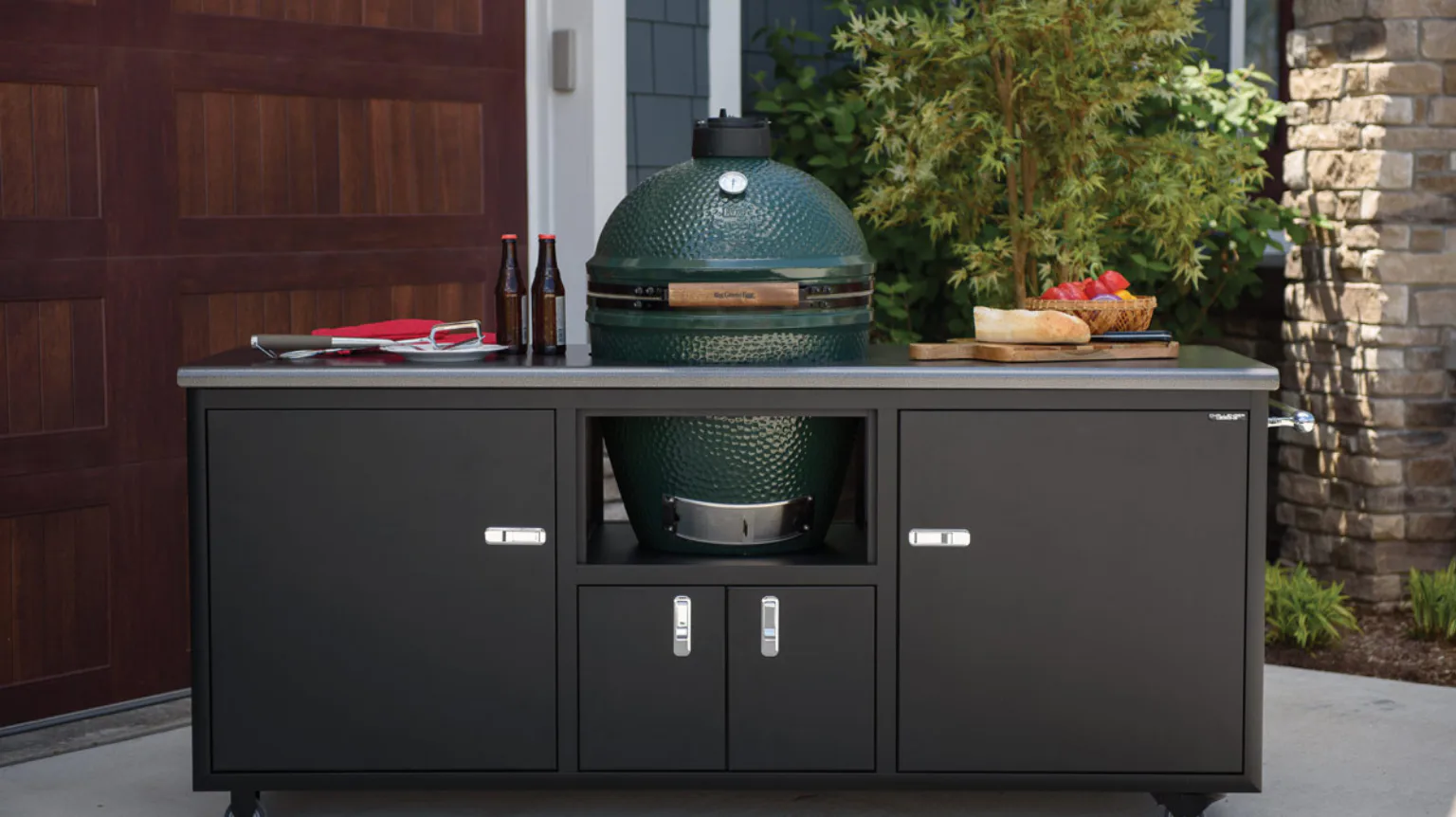 Outdoor kitchen setup with a green ceramic grill in a black cabinet. The countertop has grilling tools, bottles, bread, and fruit. Plants and a brick wall are in the background.