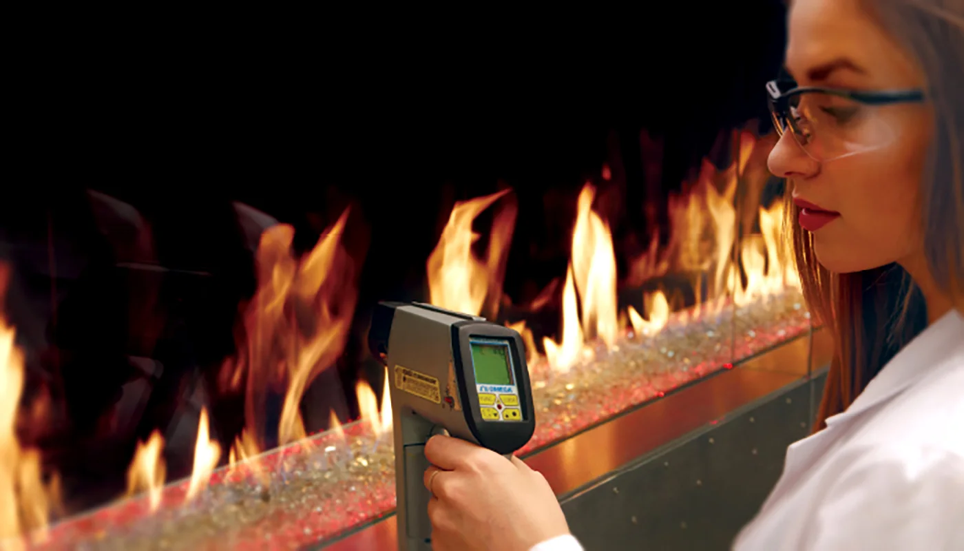 A person wearing lab goggles uses a handheld infrared thermometer to measure the temperature of a controlled fire in a laboratory setting.