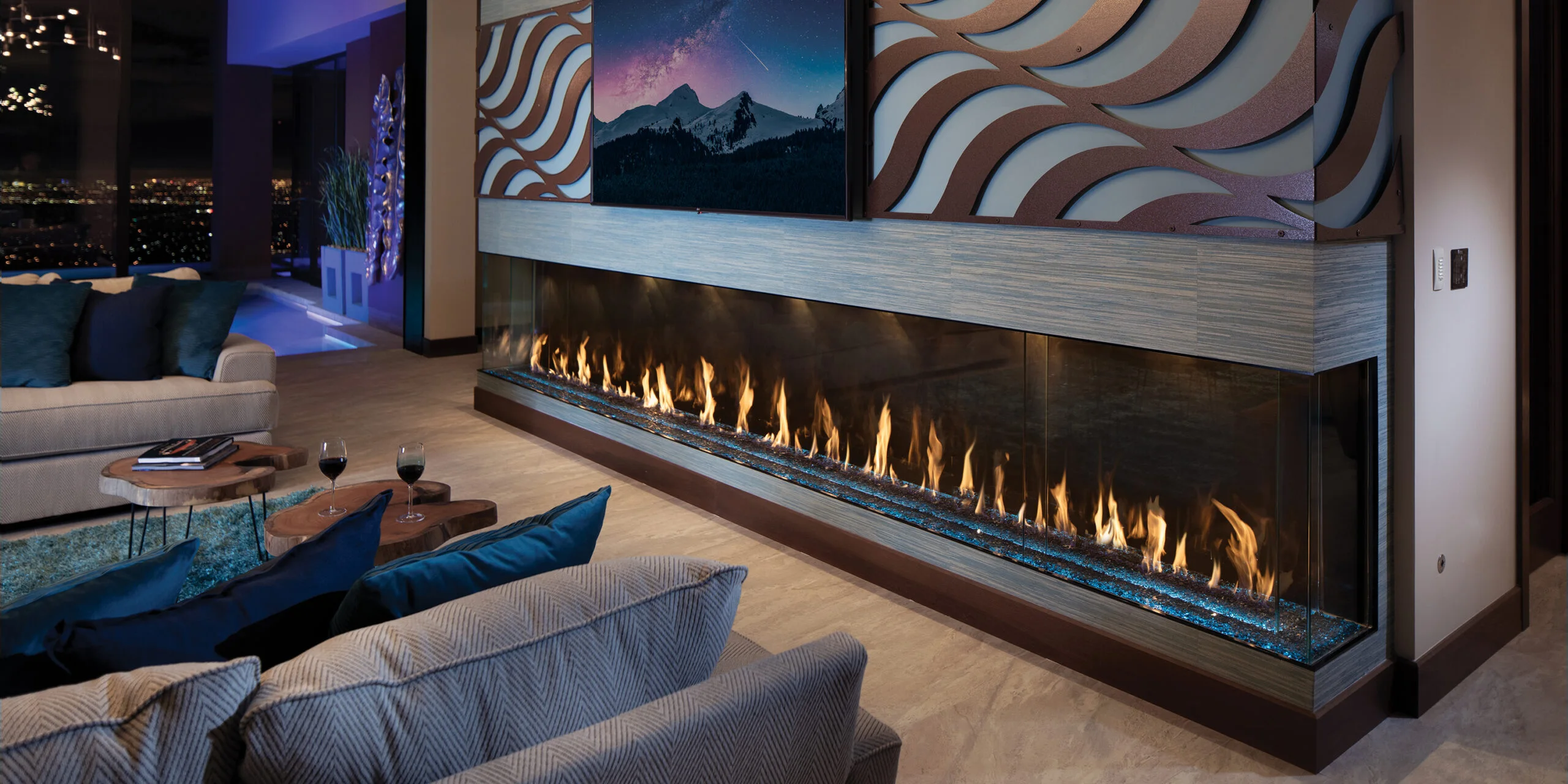 Modern living room with a long, horizontal electric fireplace under a wall-mounted TV. The room features a beige couch with blue cushions, a wood coffee table, and city views through large windows.