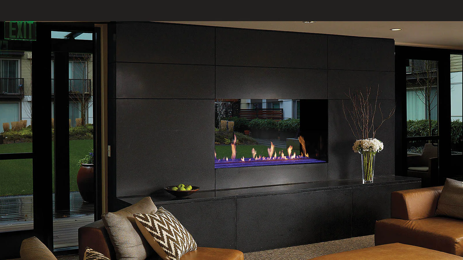 Modern living room with built-in linear fireplace showing blue and orange flames in a dark tiled wall, leather sofas, a vase of white flowers and courtyard windows