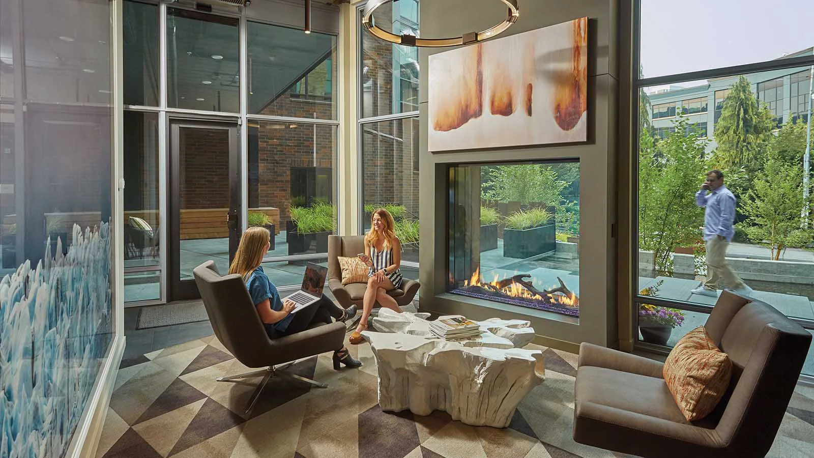 Modern glass-walled lounge with fireplace, two women conversing in armchairs (one with a laptop), sculpted coffee table, abstract wall art and greenery outside.