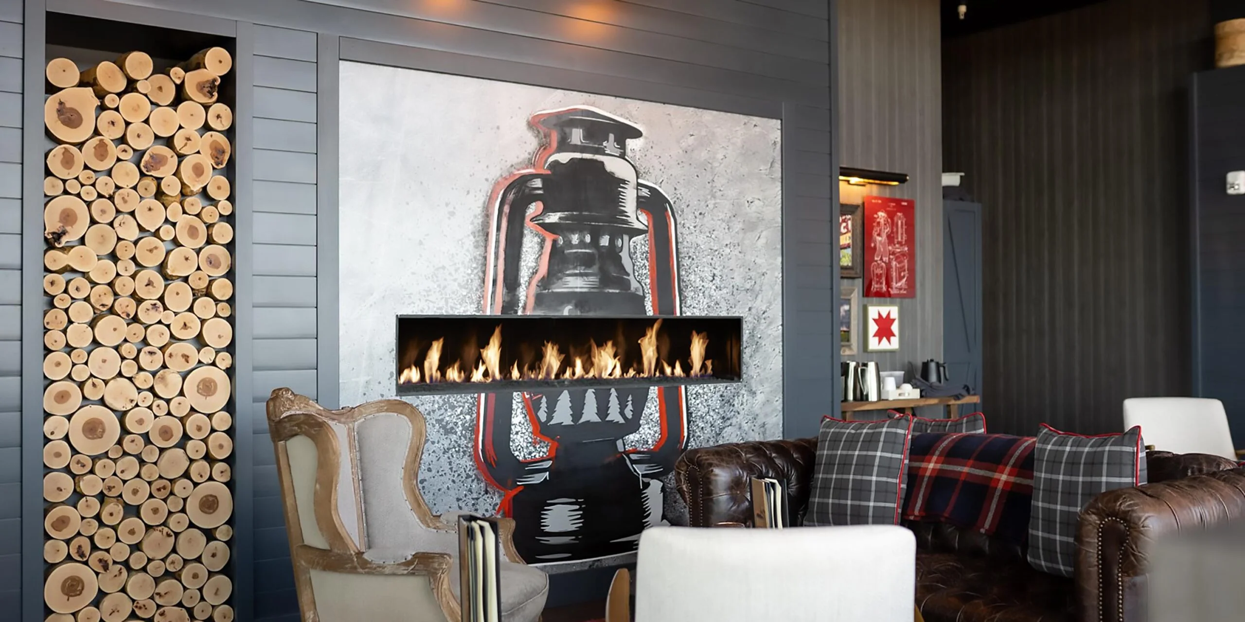 A modern lounge area featuring a wall with stacked firewood logs and a horizontal fireplace, surrounded by comfortable chairs and sofas. A vintage lantern painting is displayed above the fireplace.