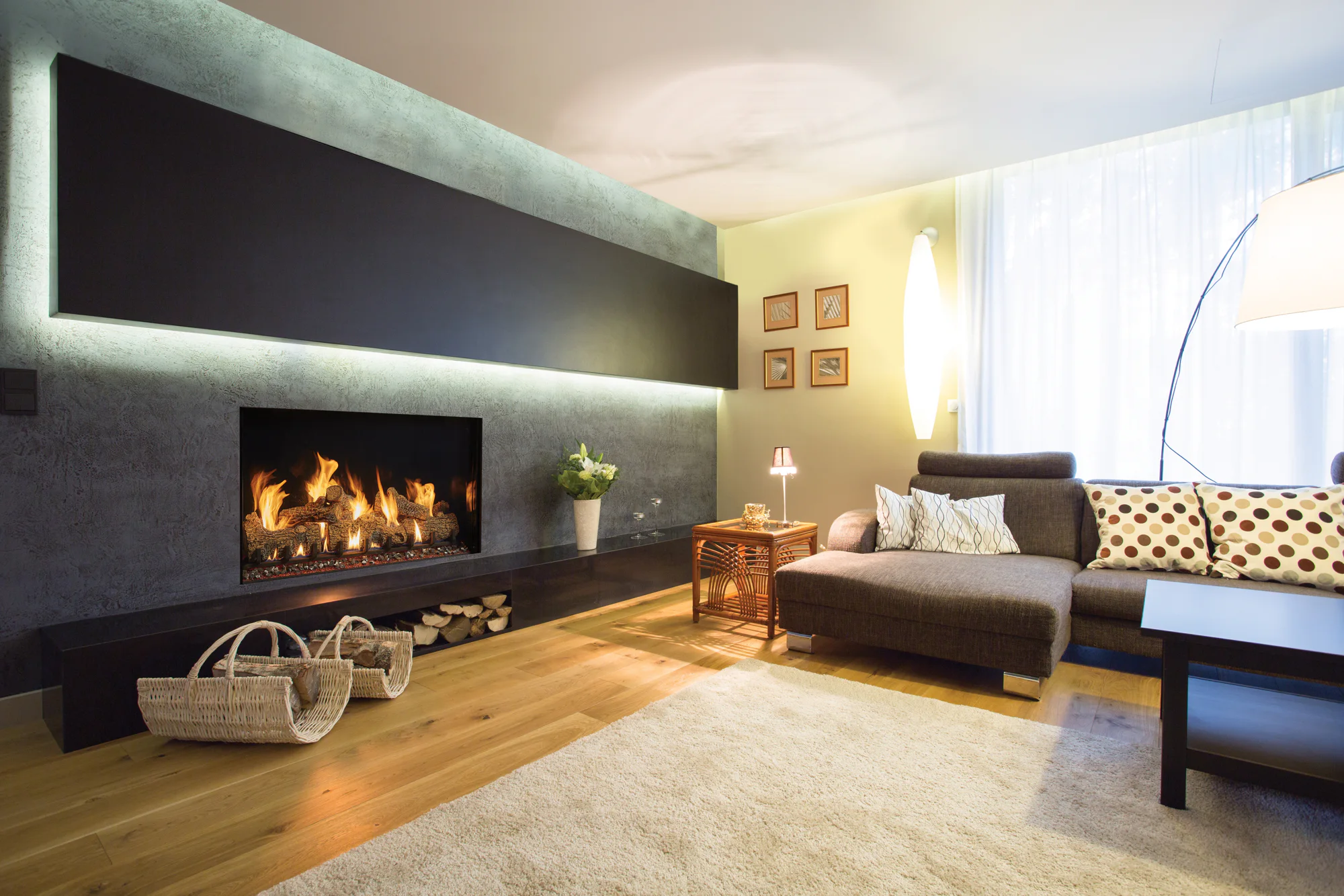 Cozy modern living room with lit fireplace, gray sectional sofa, floor lamp, wicker baskets and a large rug