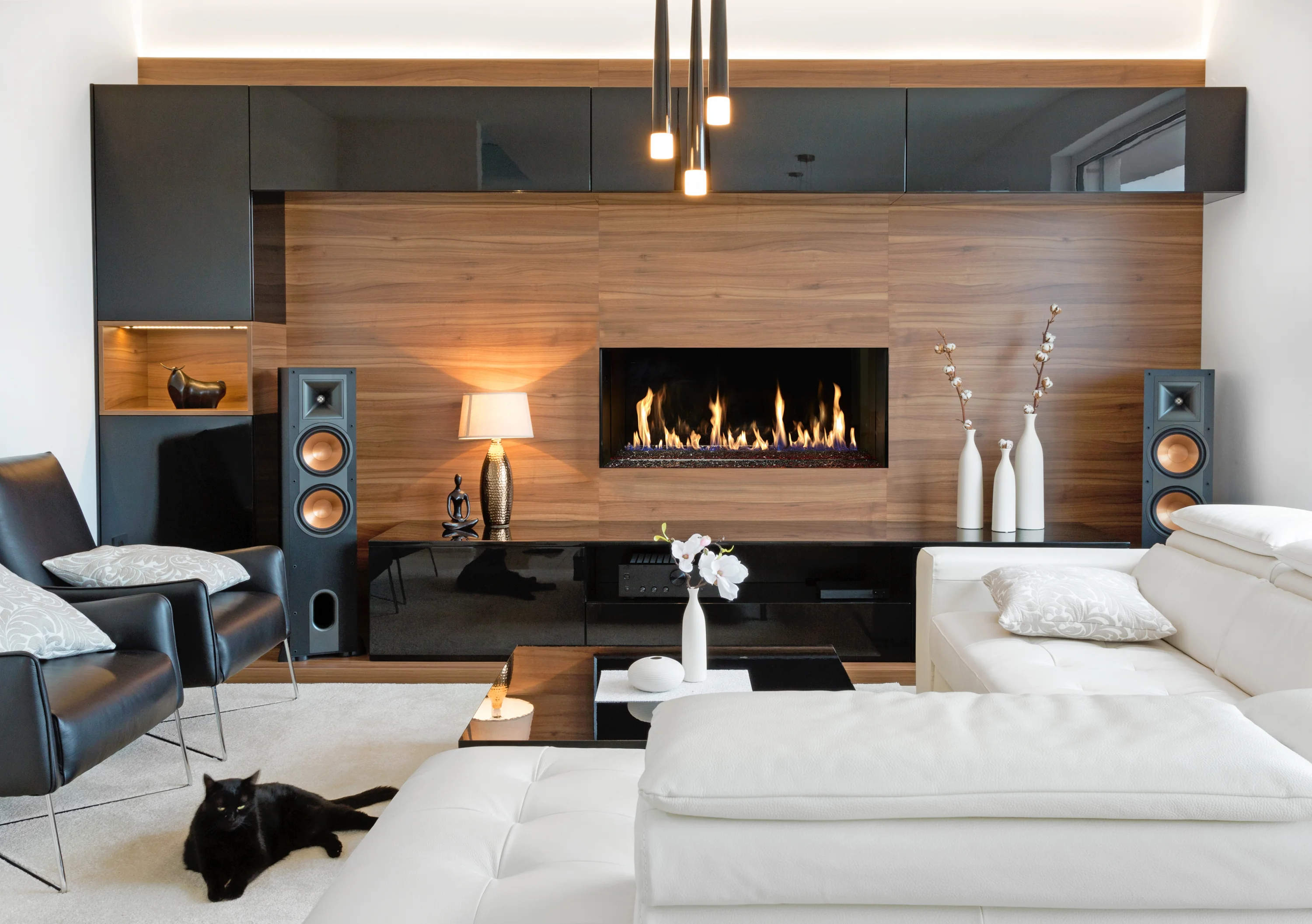 Modern living room with a sleek fireplace, black and white furniture, and a black cat on the floor. Wood paneling, decorative vases, and ambient lighting complete the cozy look.
