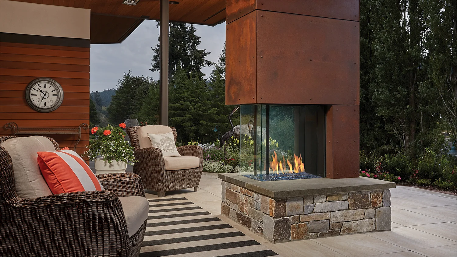 Patio with wicker chairs, striped cushions, striped rug, and a large stone fireplace. Flowers, a clock on the wall, and a scenic view of trees are visible in the background.