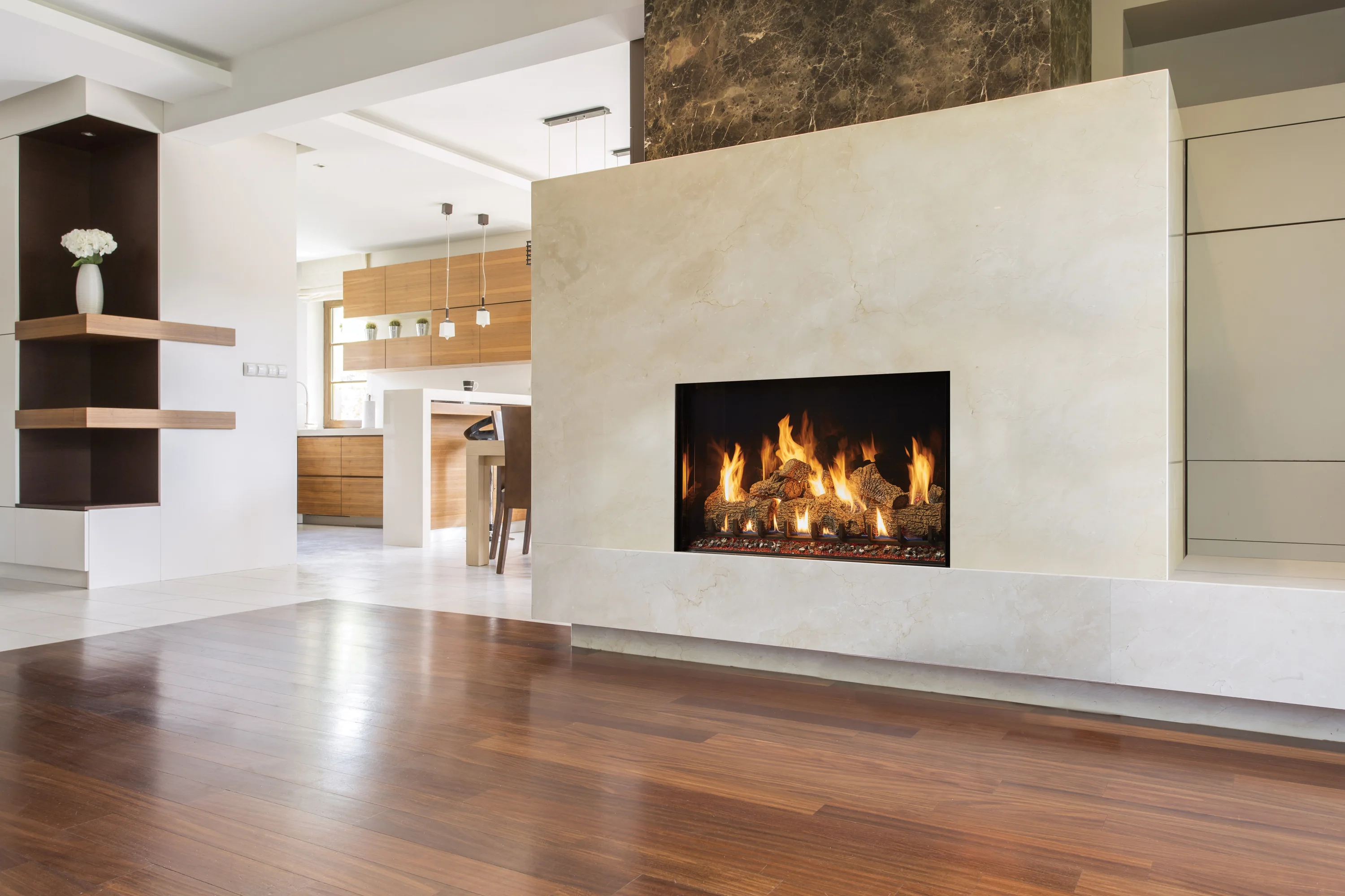 Open modern living room with marble-framed gas fireplace, dark hardwood floors, and view into a wood-finish kitchen with pendant lights and a white vase.