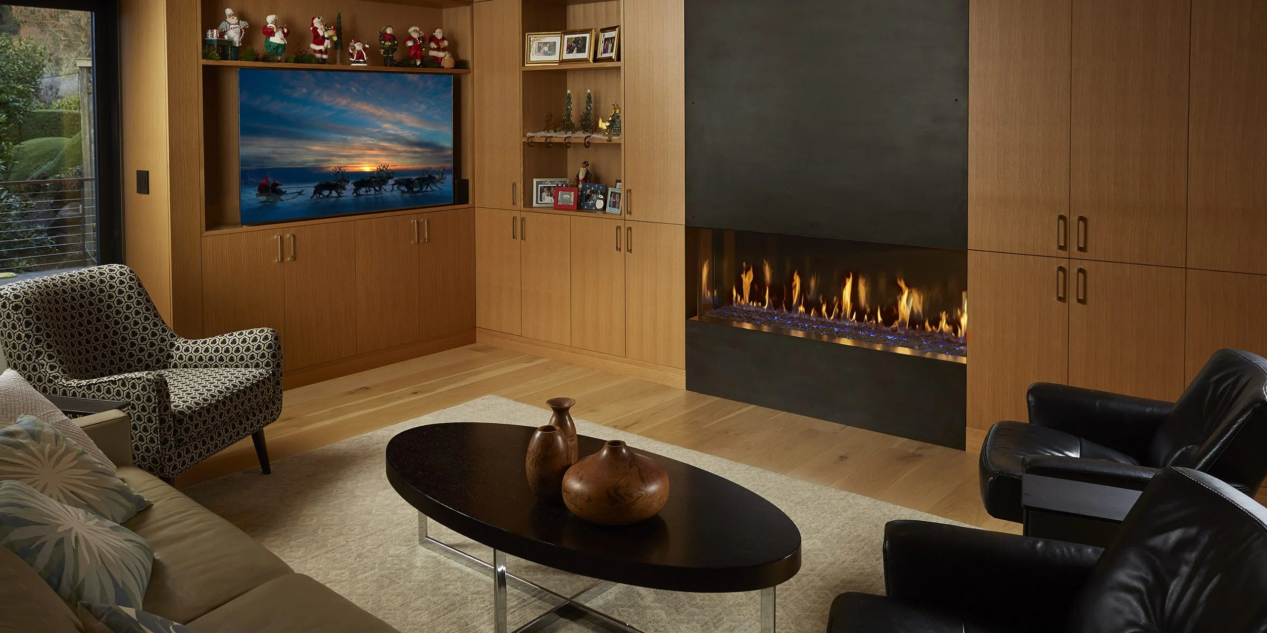 Modern living room with wooden cabinets, a fireplace, and a TV displaying a winter scene. There are Christmas decorations, framed photos, an oval coffee table with two vases, and various seating.