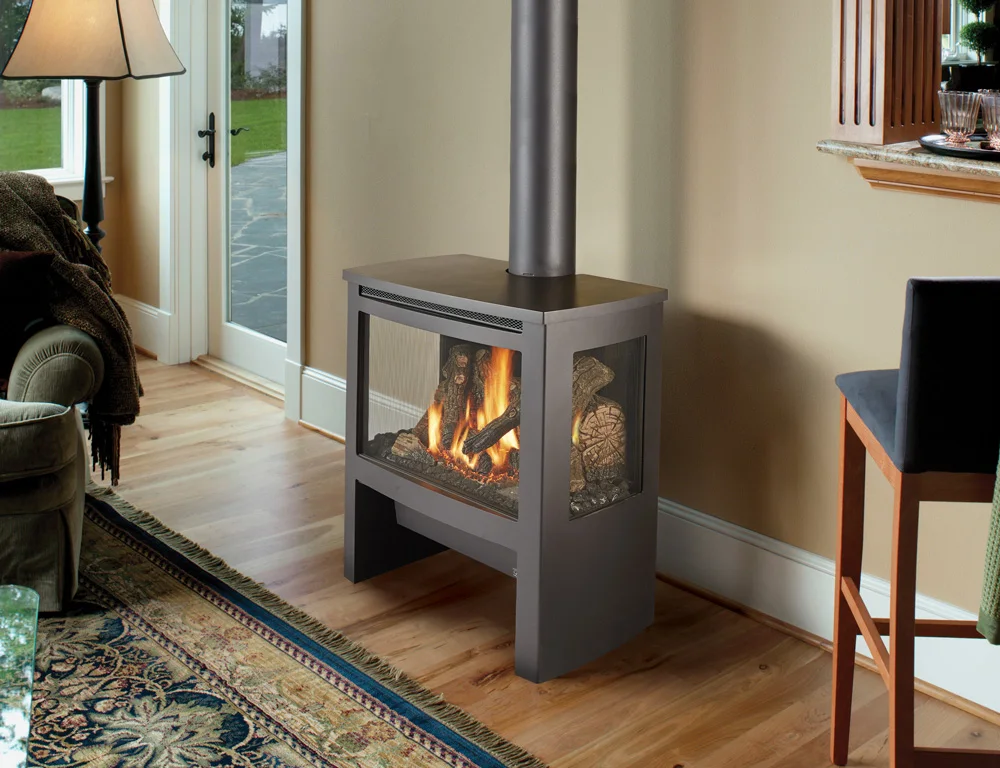 Modern living room with hardwood flooring, a patterned rug, and a corner wood stove with visible flames. A large window with French doors opens to an outdoor patio.
