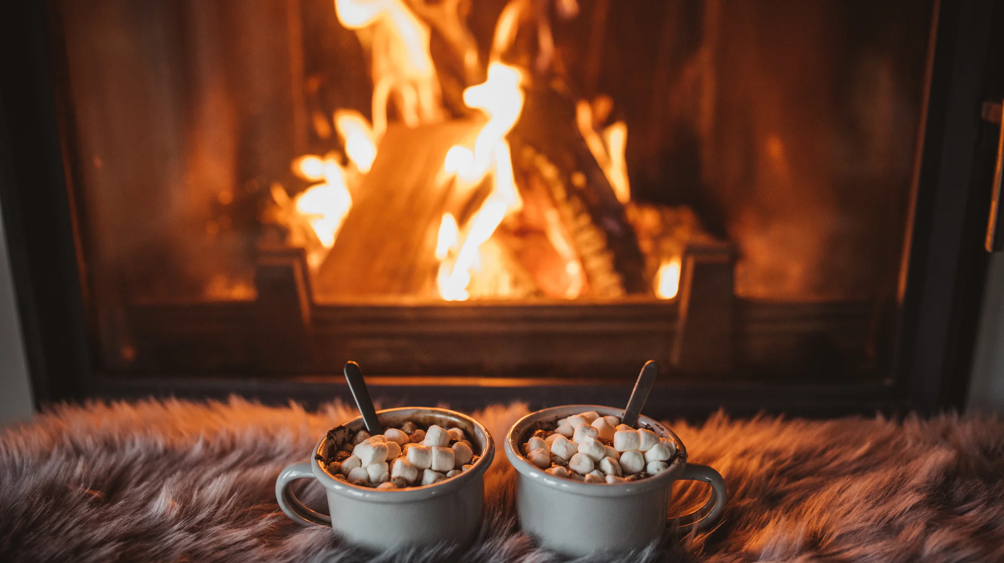 Two mugs of hot chocolate topped with marshmallows on a fuzzy surface in front of a burning fireplace, creating a cozy and warm atmosphere.