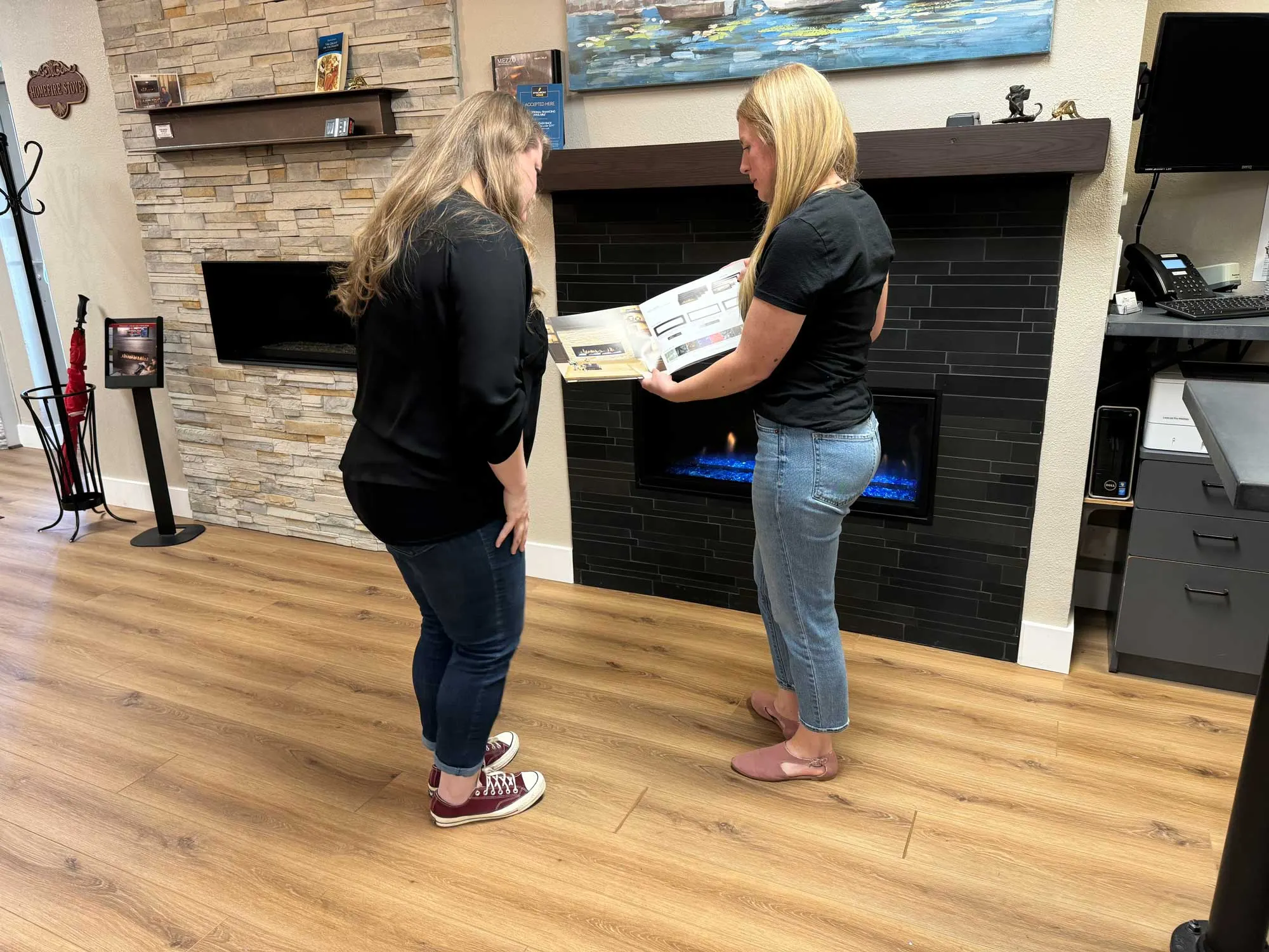 Two people in a fireplace showroom discuss a brochure. One person shows the brochure to the other, while an electric fireplace with a blue flame glows in the background.