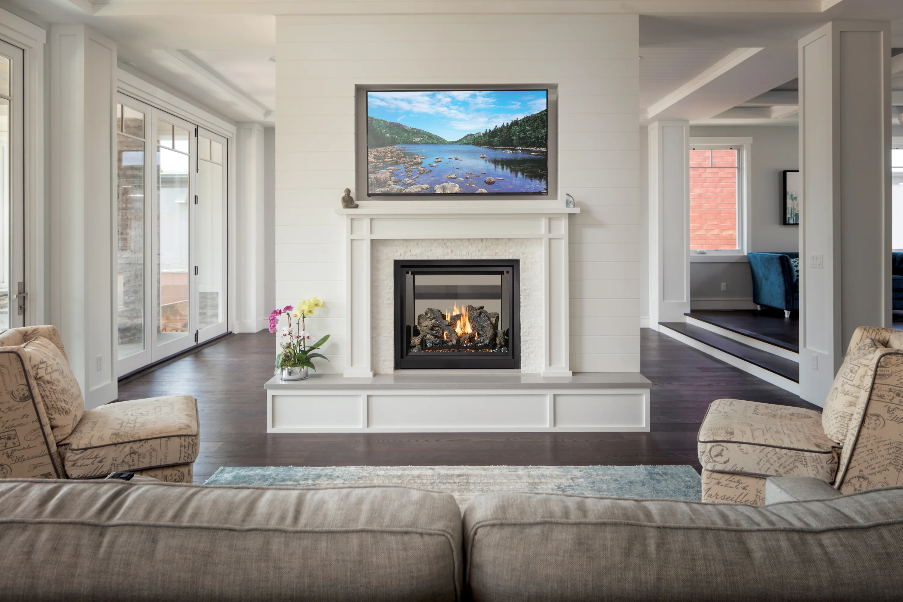 Bright modern living room centered on a white fireplace with a burning gas fire, TV above showing a lake scene, sofas and large windows