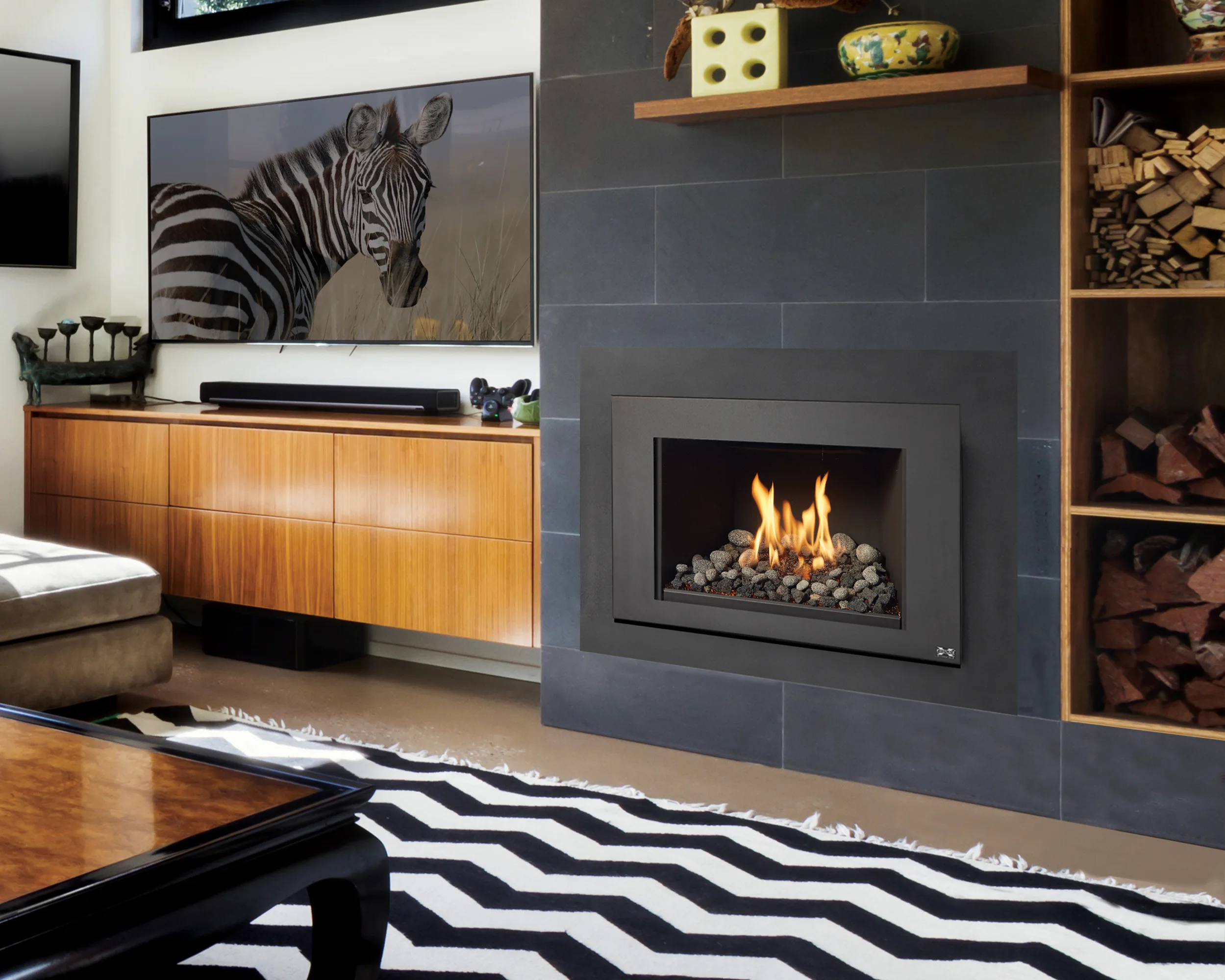 Living room with a modern fireplace, a zebra artwork on the wall, a wooden sideboard, and a black and white zigzag rug. Firewood is stacked on shelves to the right.
