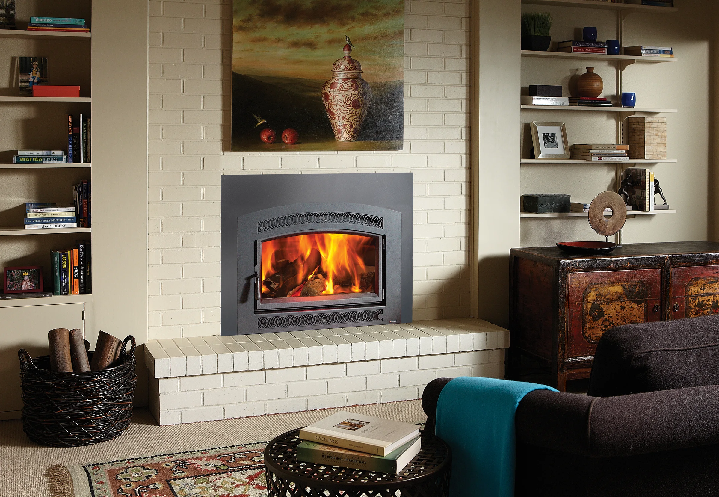 Wood-burning fireplace in white brick with painting above, built-in shelves of books and decor, basket of logs, rug and dark sofa with teal throw