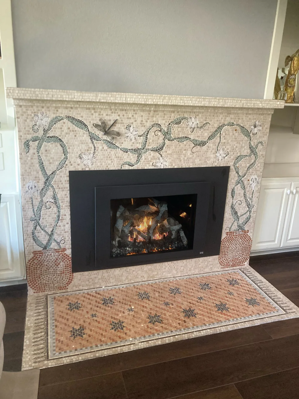 A fire burns inside a black metal fireplace, which is adorned with a detailed mosaic, featuring green vines, white flowers, and a dragonfly. The hearth is also covered in patterned mosaic tiles.
