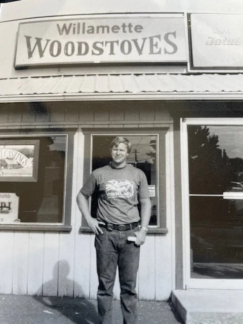 Man standing in front of a building with a sign that reads Willamette Woodstoves. He is wearing a t-shirt and jeans, and holding an object in his right hand.
