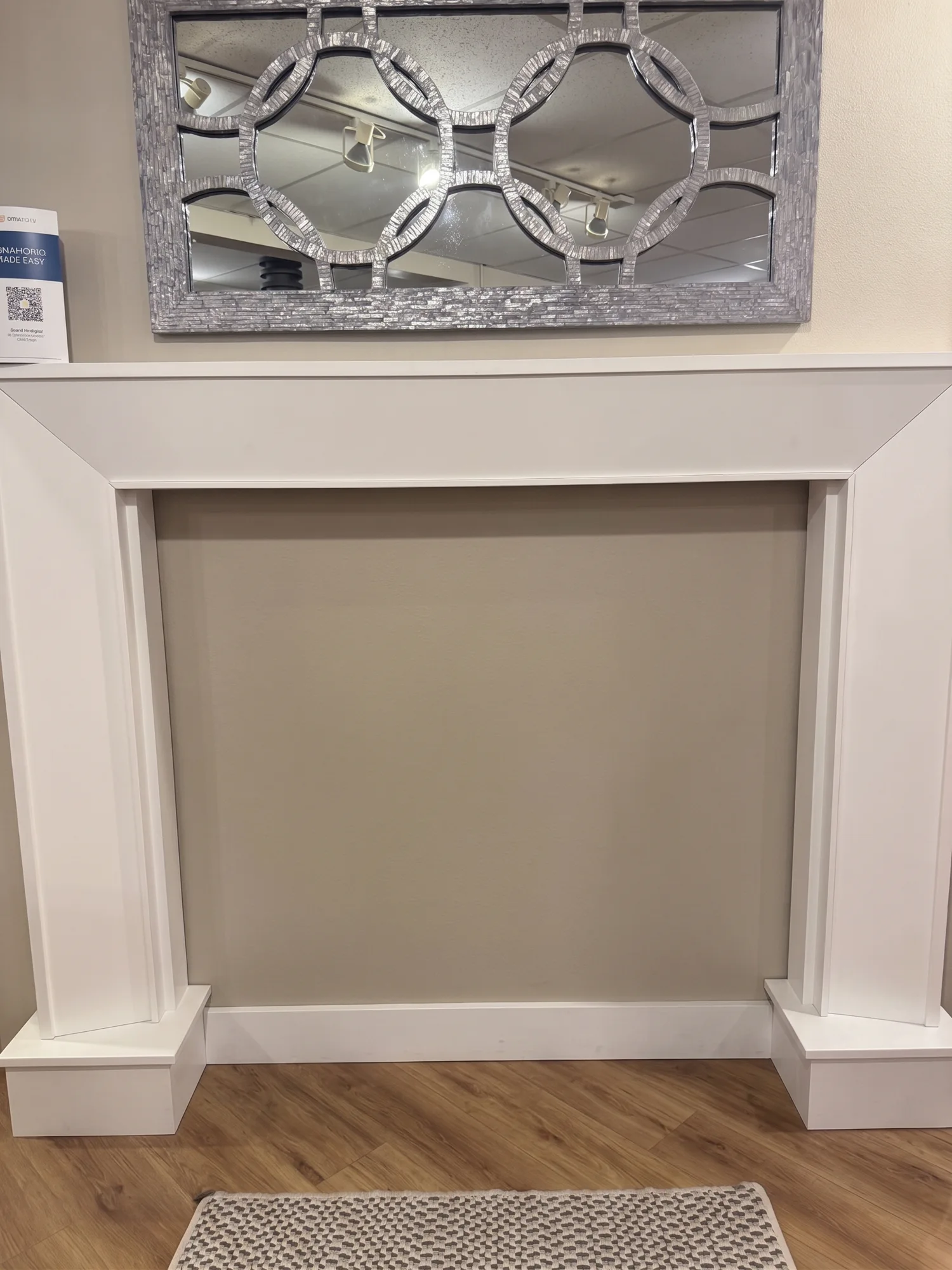 Silver decorative mirror above a white mantel on a beige wall, with wood floor and a small rug
