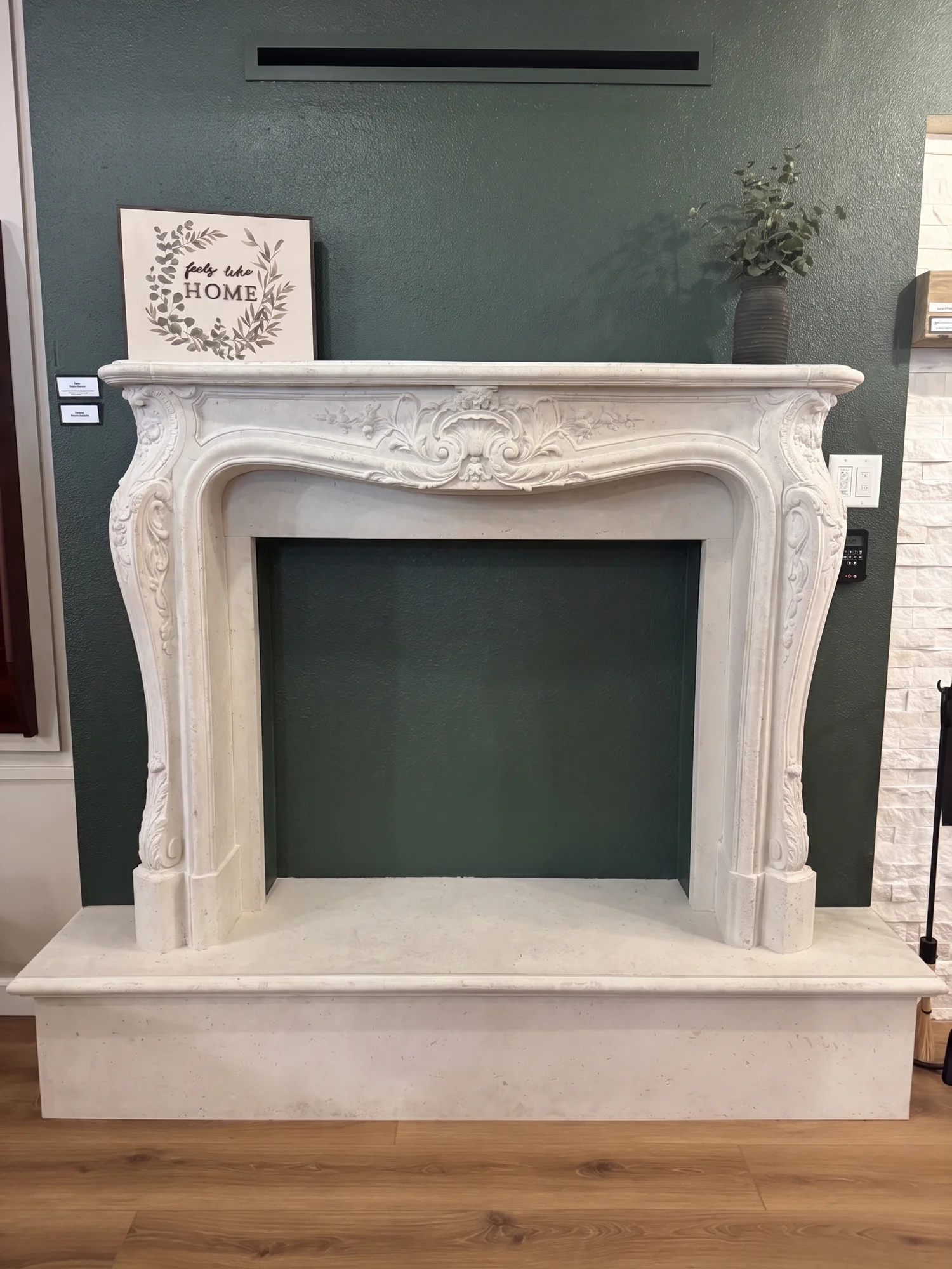 Ornate carved white fireplace mantel against a dark green wall with a framed sign reading 'feels like HOME' and a vase of greenery