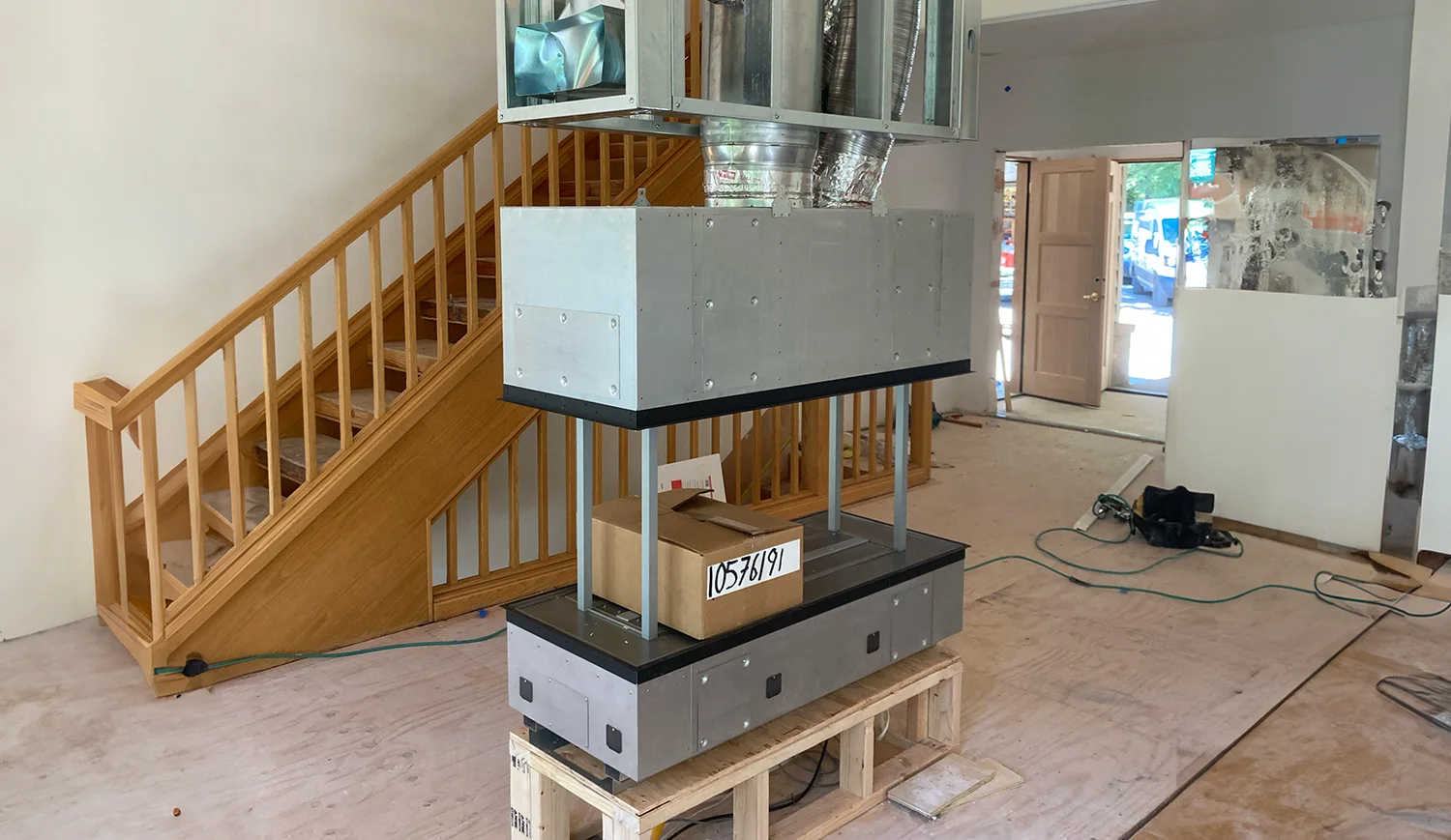 Unfinished interior with a large metal HVAC/fireplace unit mounted on a wooden pallet in the center, wood staircase to left, open doorway and tools to right.