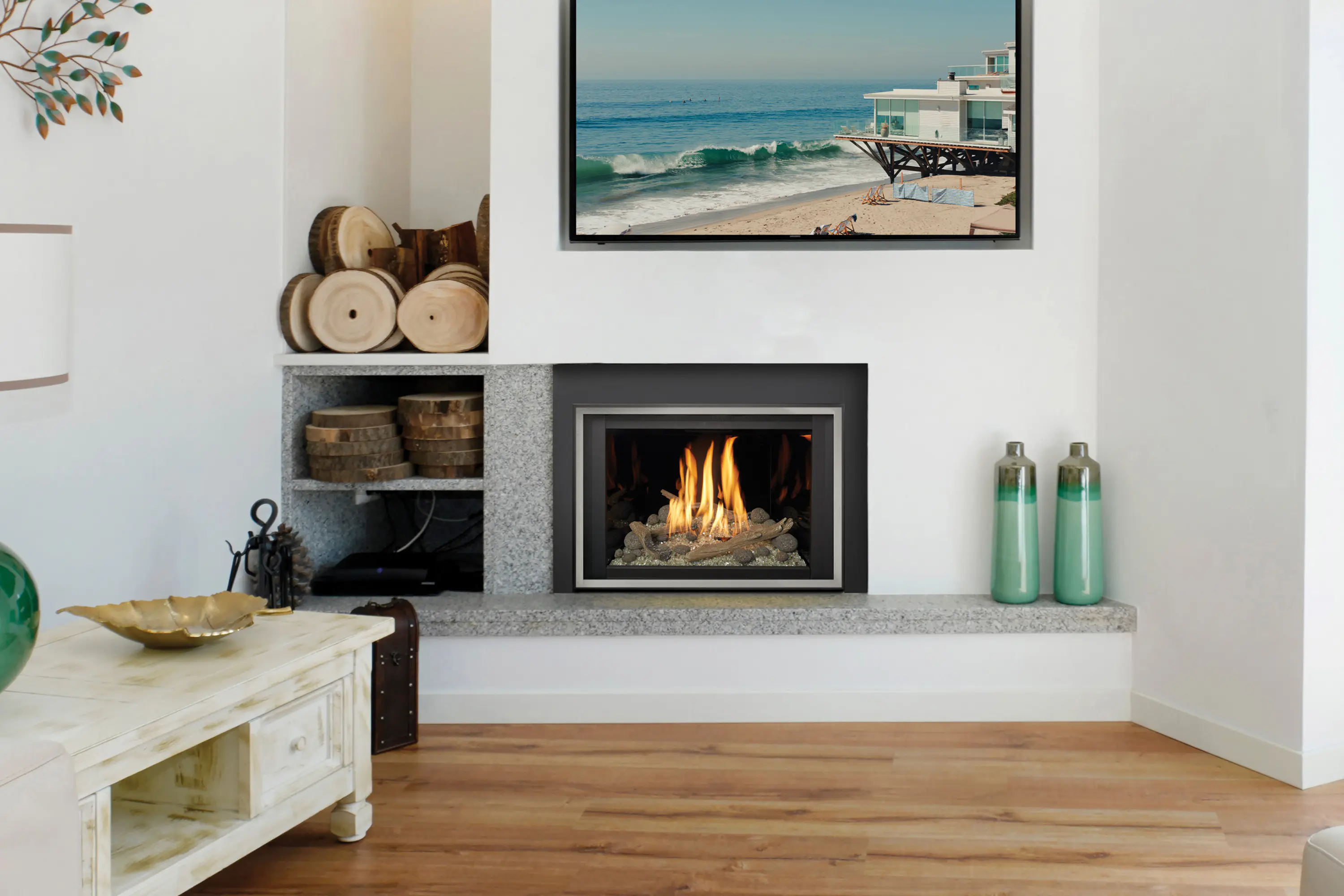 Modern living room with a fireplace, stacked logs, and a beach photo above. A white table with a gold bowl and green vase, and teal vases on the right. Light wood flooring.