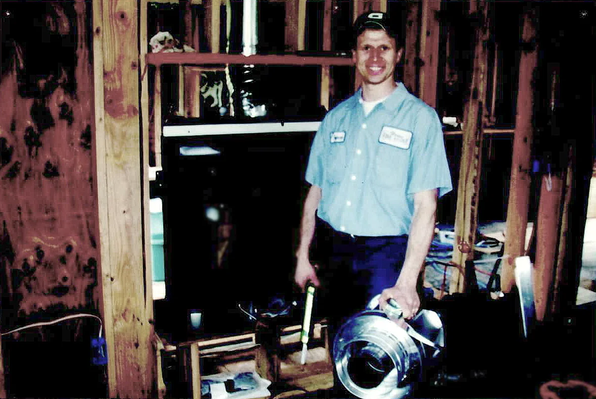 Man in a blue work uniform and cap stands in a partially constructed room holding a metal duct and a tool. Wooden beams and construction materials are visible around him.