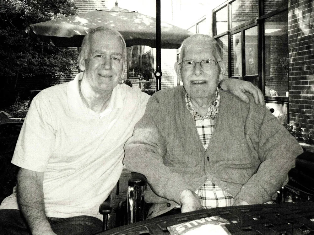 Two older adults sitting outdoors at a table, one with an arm around the other. They are both smiling. A large umbrella and brick building are in the background.