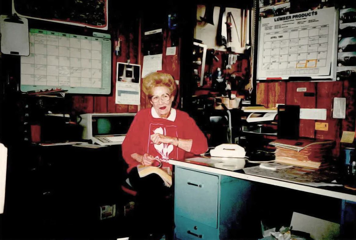 Elderly woman in a red sweater sits in a cluttered office with a calendar on the wall. She holds a pen and notebook, surrounded by office supplies and equipment.