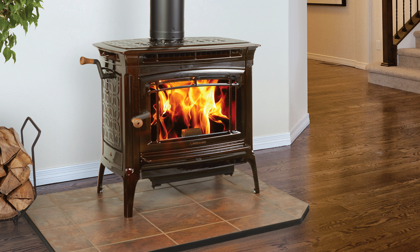 A wood-burning stove with a roaring fire sits on a tile platform next to a metal log holder with stacked wood. The stove is located in a room with hardwood floors and stairs.